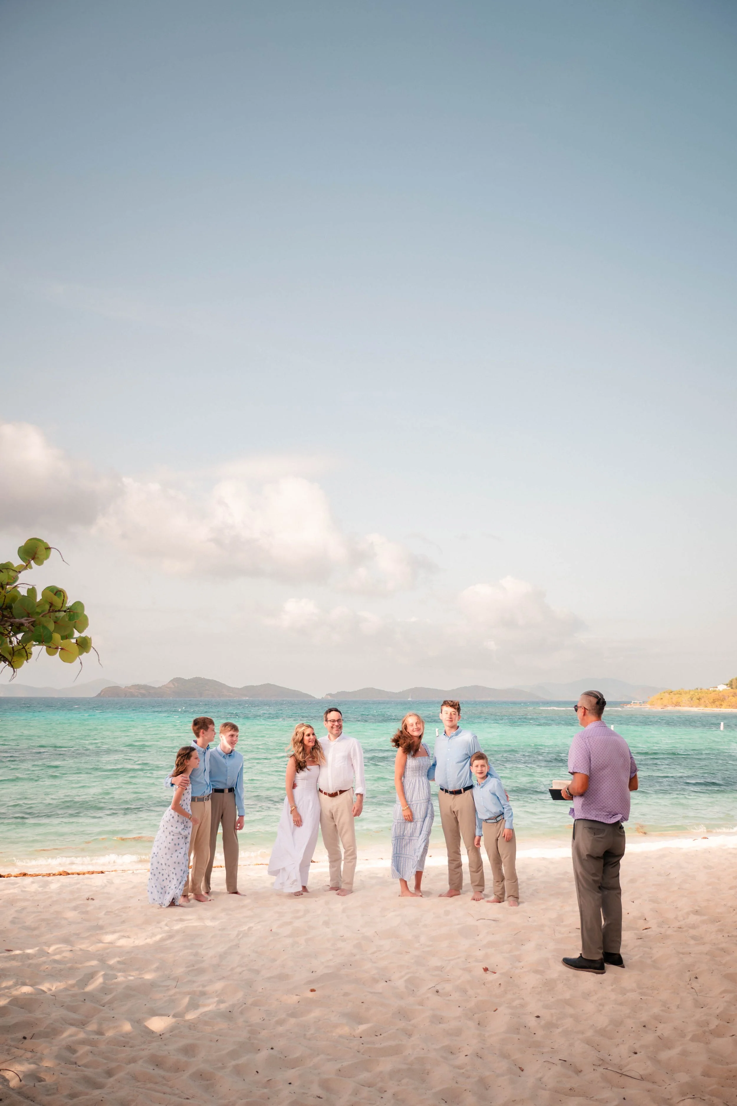 A group of people is on a sandy beach near turquoise ocean water, with mountains and cloudy sky in the background, during a daytime celebration or gathering led by a man with a clipboard.