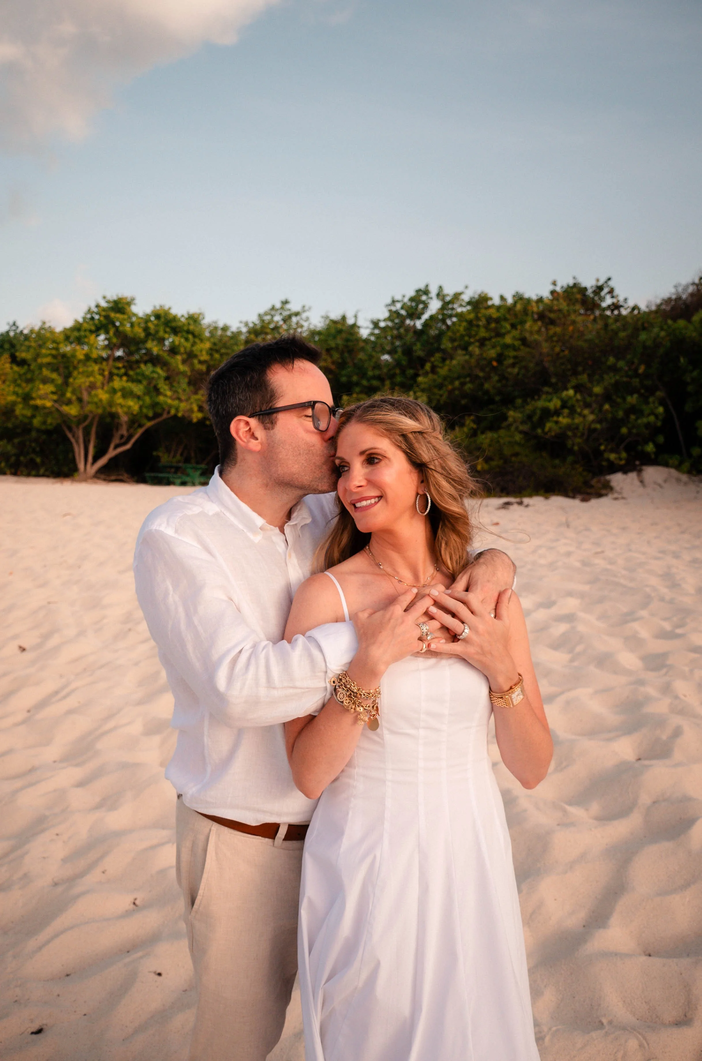 A couple standing on a sandy beach, embracing and smiling. The man is kissing the woman's forehead, and both are dressed in light-colored clothing. There are green bushes and trees in the background, under a partly cloudy sky.