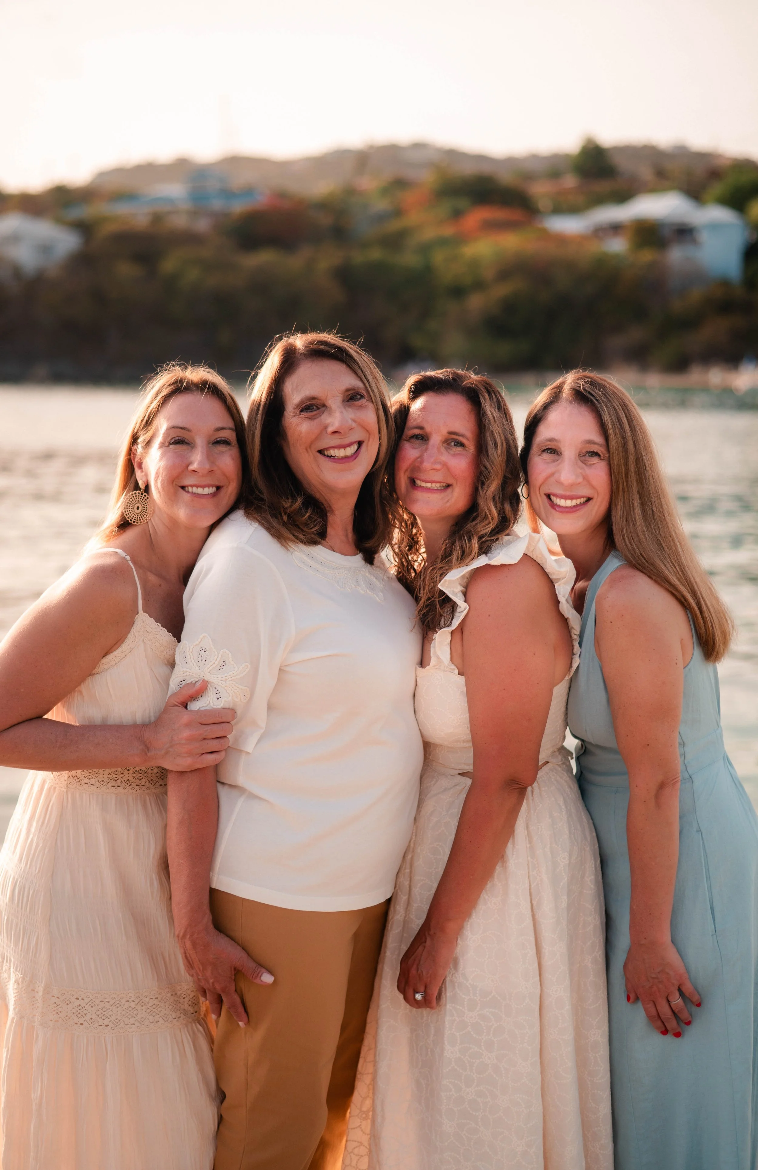 Four women smiling and standing close together outdoors with a body of water and houses on a hill in the background.