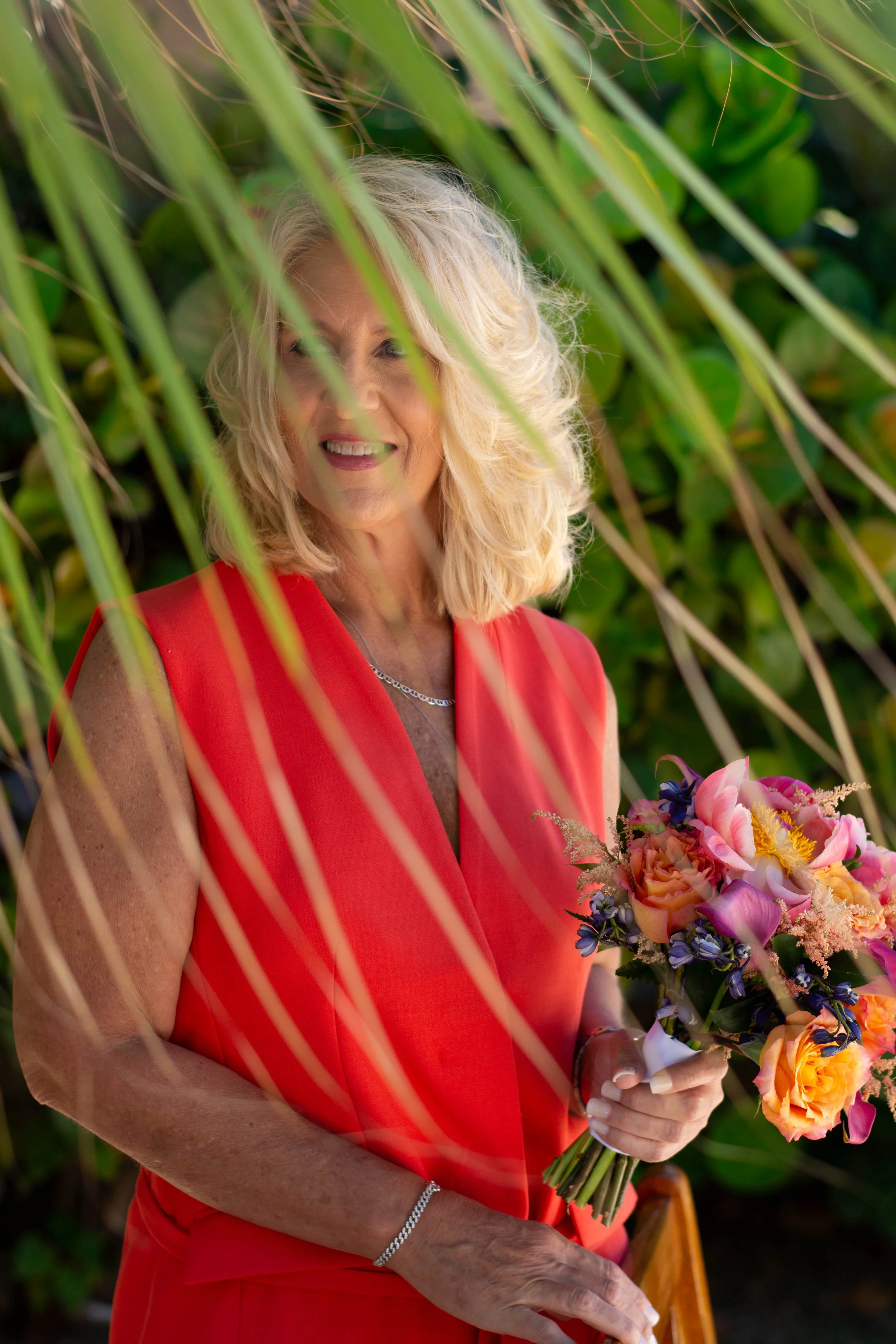 A woman with blonde hair and light skin wearing a red dress, holding a bouquet of colorful flowers, standing outdoors near greenery.