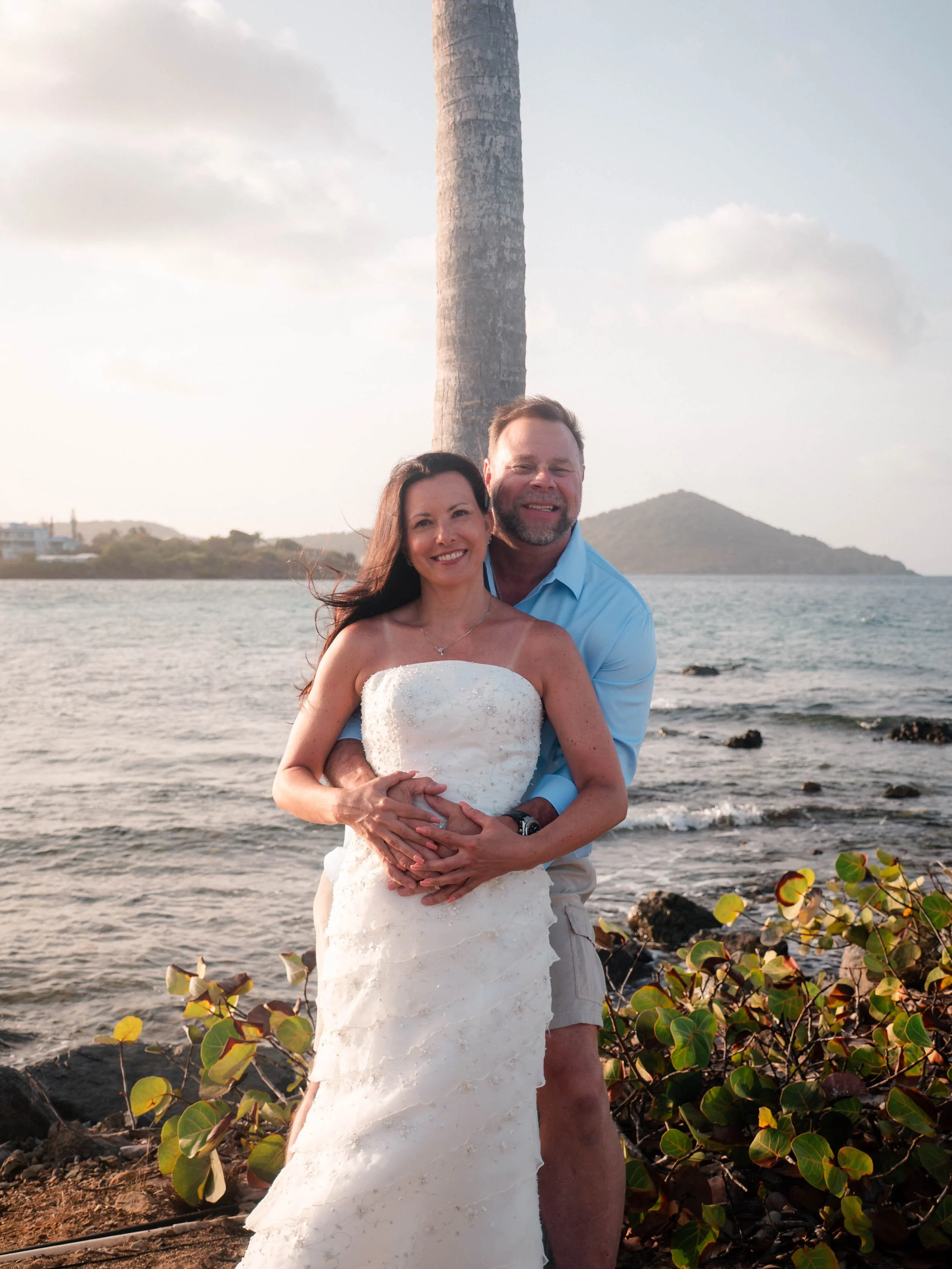 A smiling couple in wedding attire standing on a beach with water, a distant island, and a lighthouse in the background, holding a wedding cake in front of them.