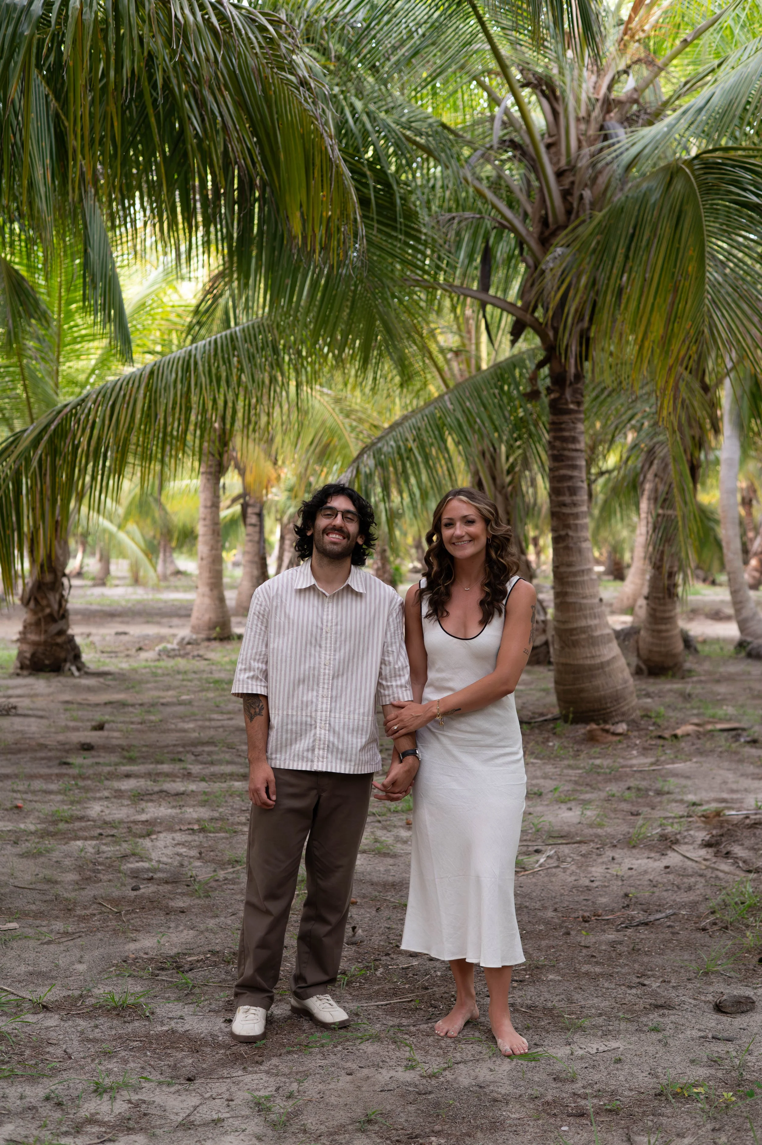 A man and woman standing together in a tropical outdoor setting with tall palm trees and a dirt ground. The man has curly hair, glasses, a striped shirt, and brown pants, while the woman has wavy hair, a white dress, and no shoes. They are smiling an