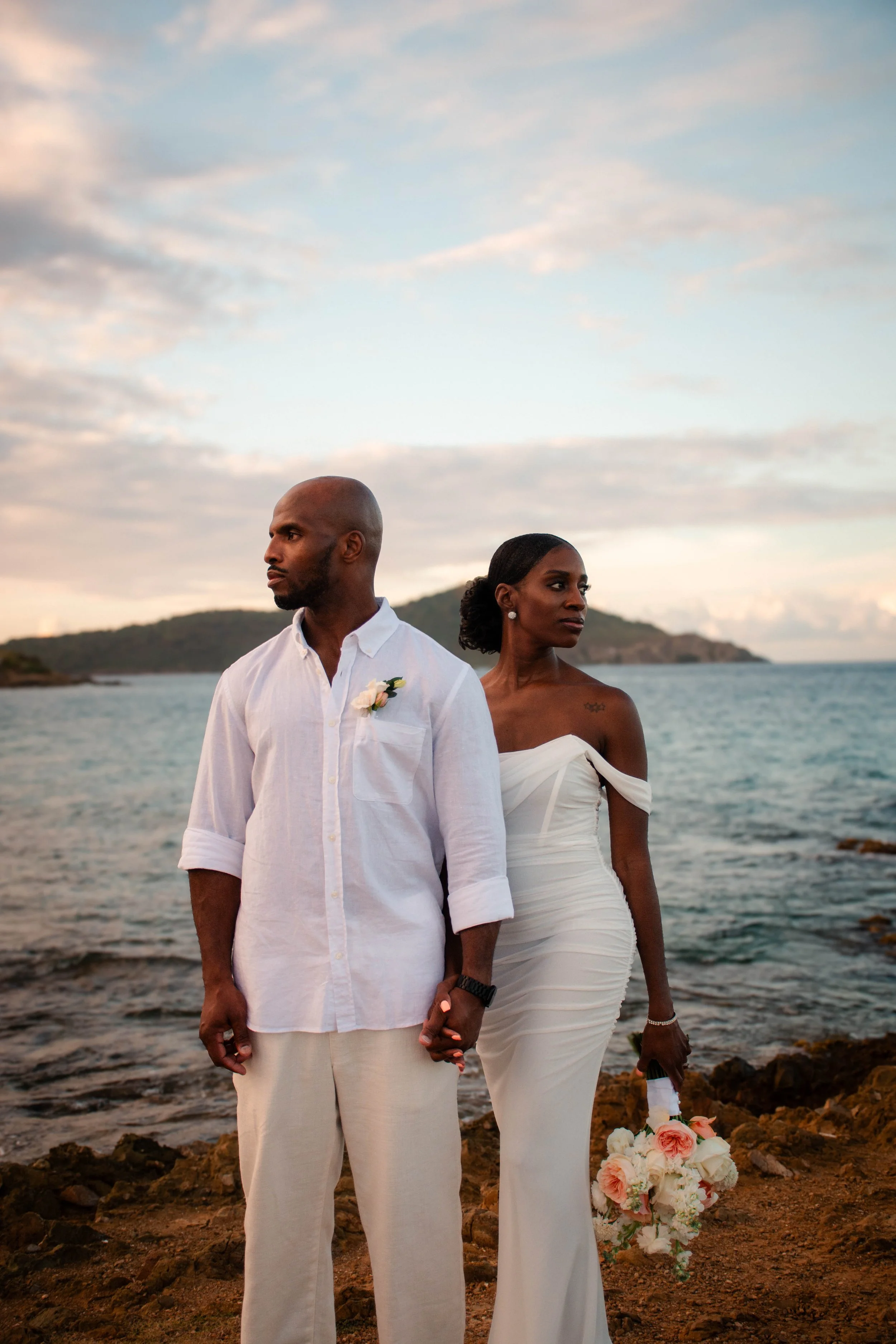 A couple dressed in white standing on a rocky shoreline near the water, holding hands, with the man looking to the side and the woman looking ahead. The woman holds a bouquet of flowers, and the woman has a tattoo on her shoulder. The sky is partly c