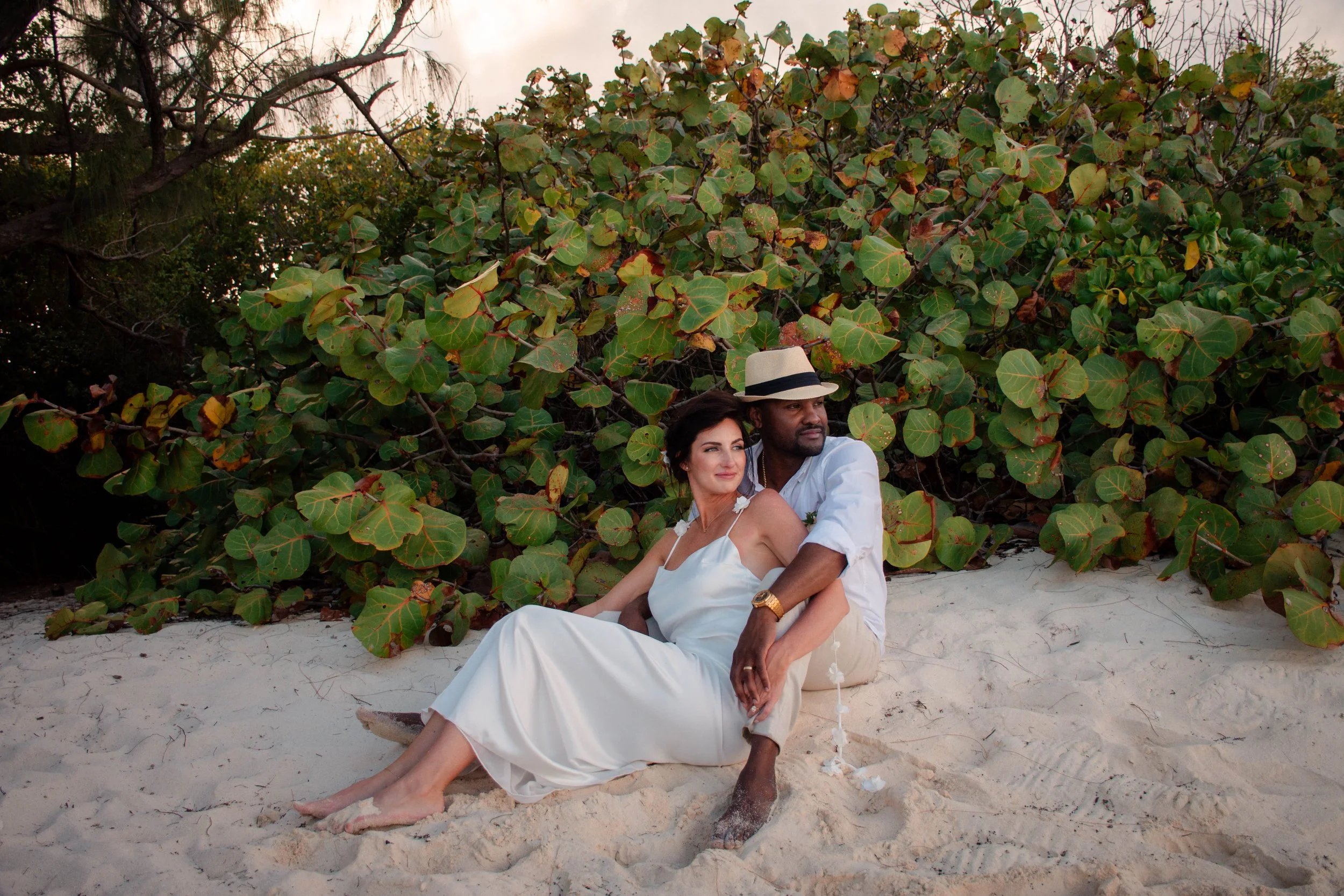 A couple sitting on a sandy beach in front of large green foliage. The woman is wearing a white dress and the man is wearing a white shirt with a beige hat. They are close with the woman's head against the man's shoulder, both looking relaxed.