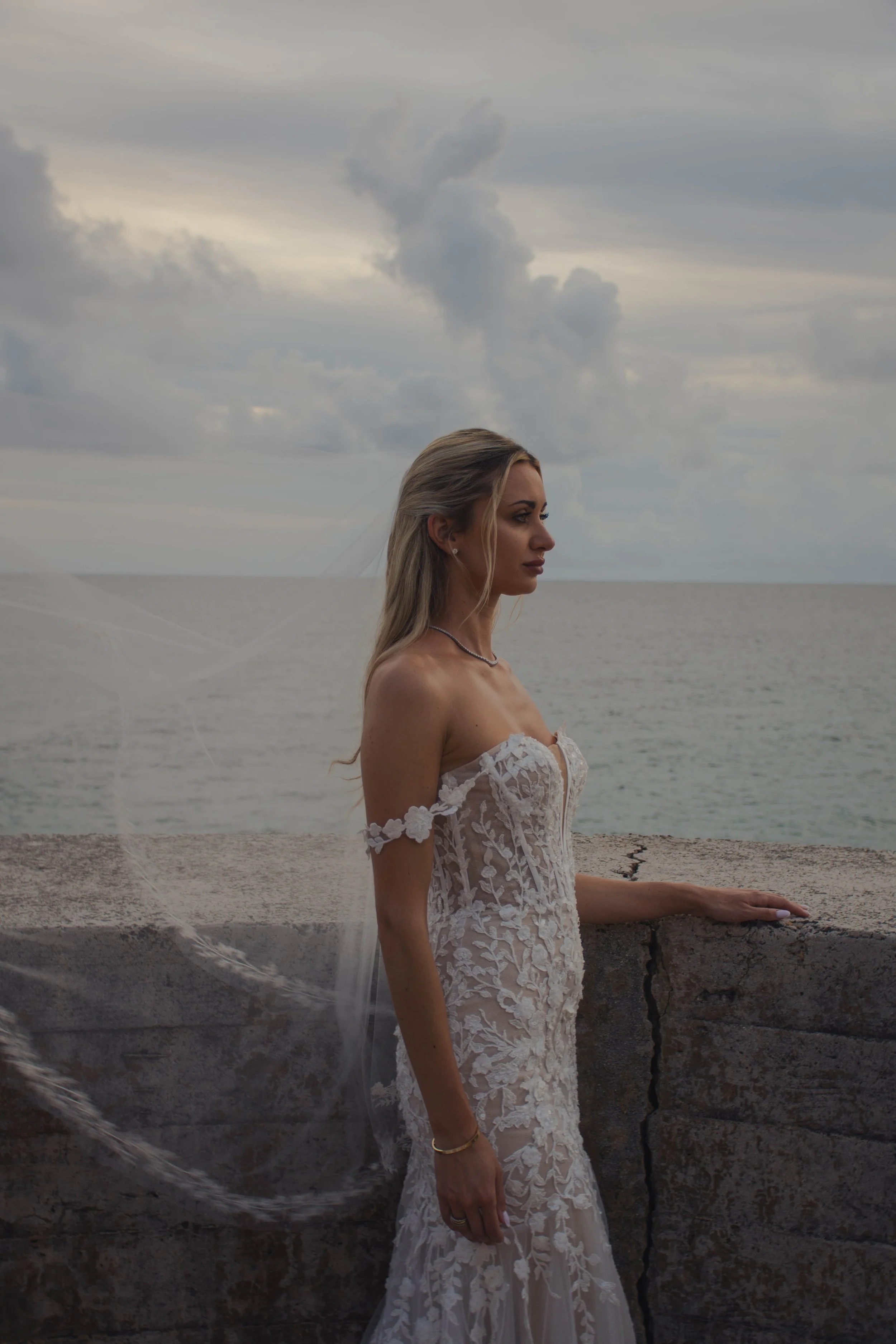 A woman in a white lace off-shoulder wedding dress standing by a stone wall at the beach on a cloudy day, with the ocean and sky in the background.