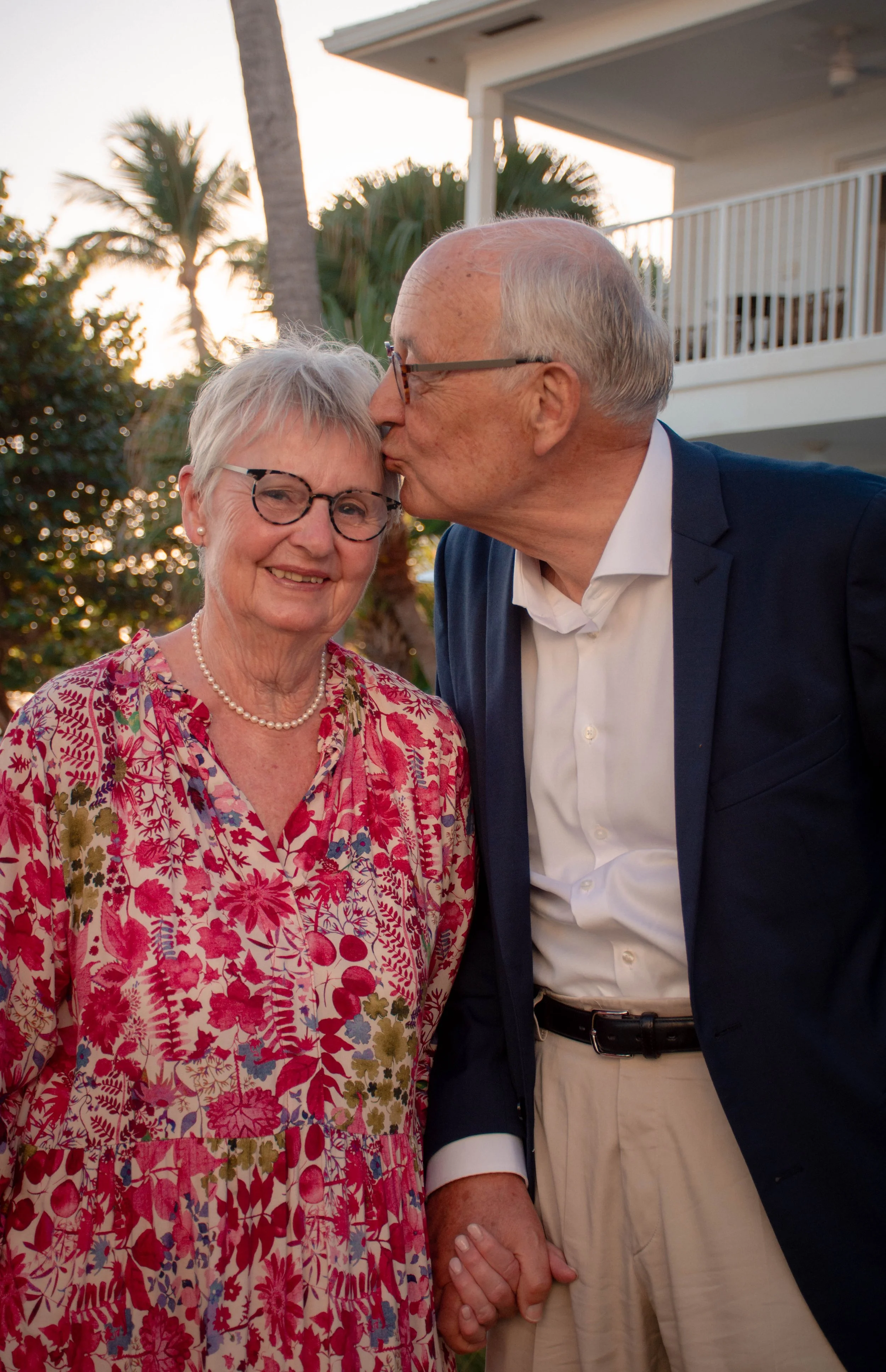 An elderly couple dressed in formal attire, holding hands, with the man giving the woman a kiss on the forehead outdoors during sunset.