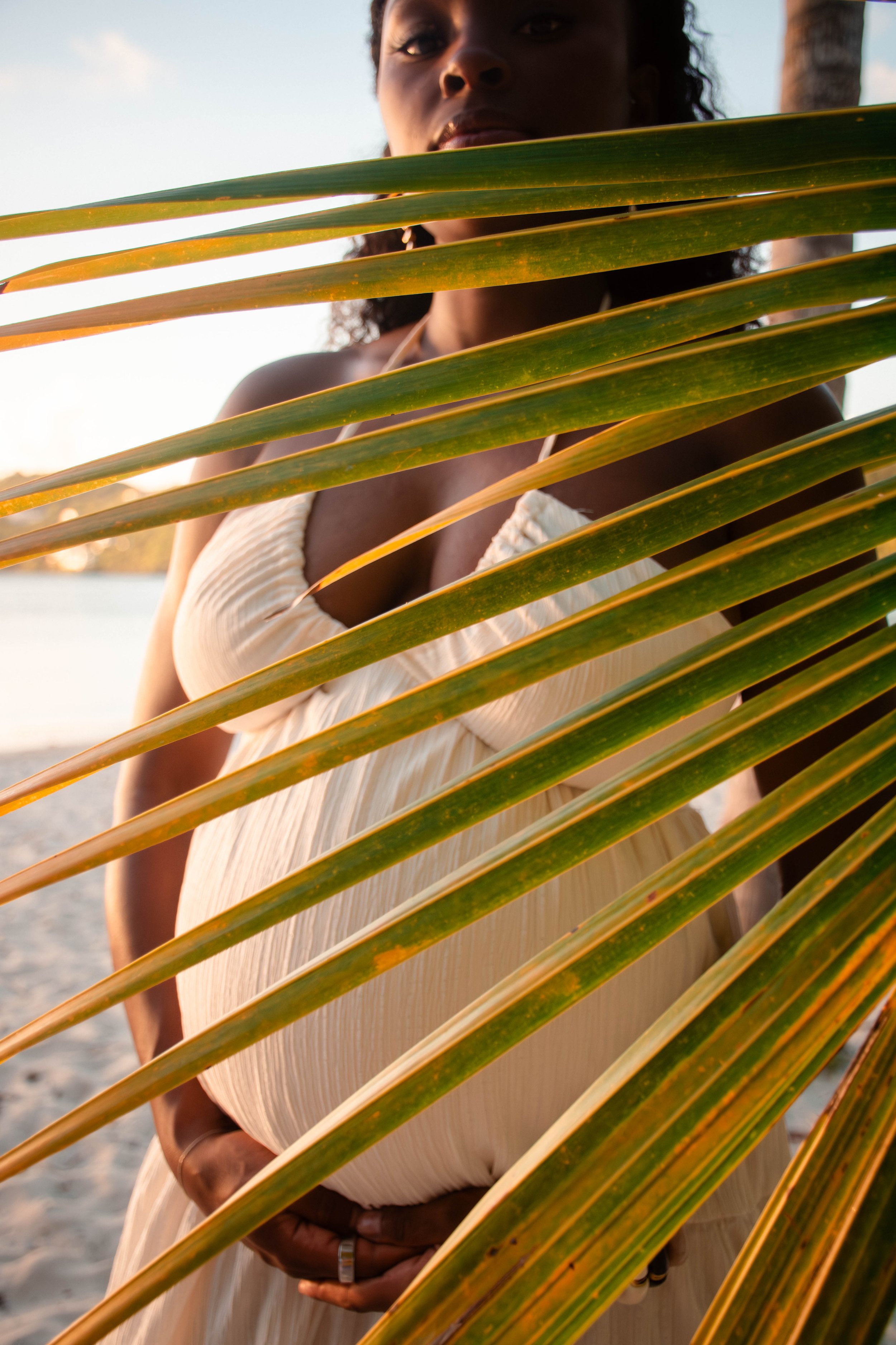 A person with dark skin and curly hair standing behind palm leaves, on a beach at sunset, wearing a light-colored dress.