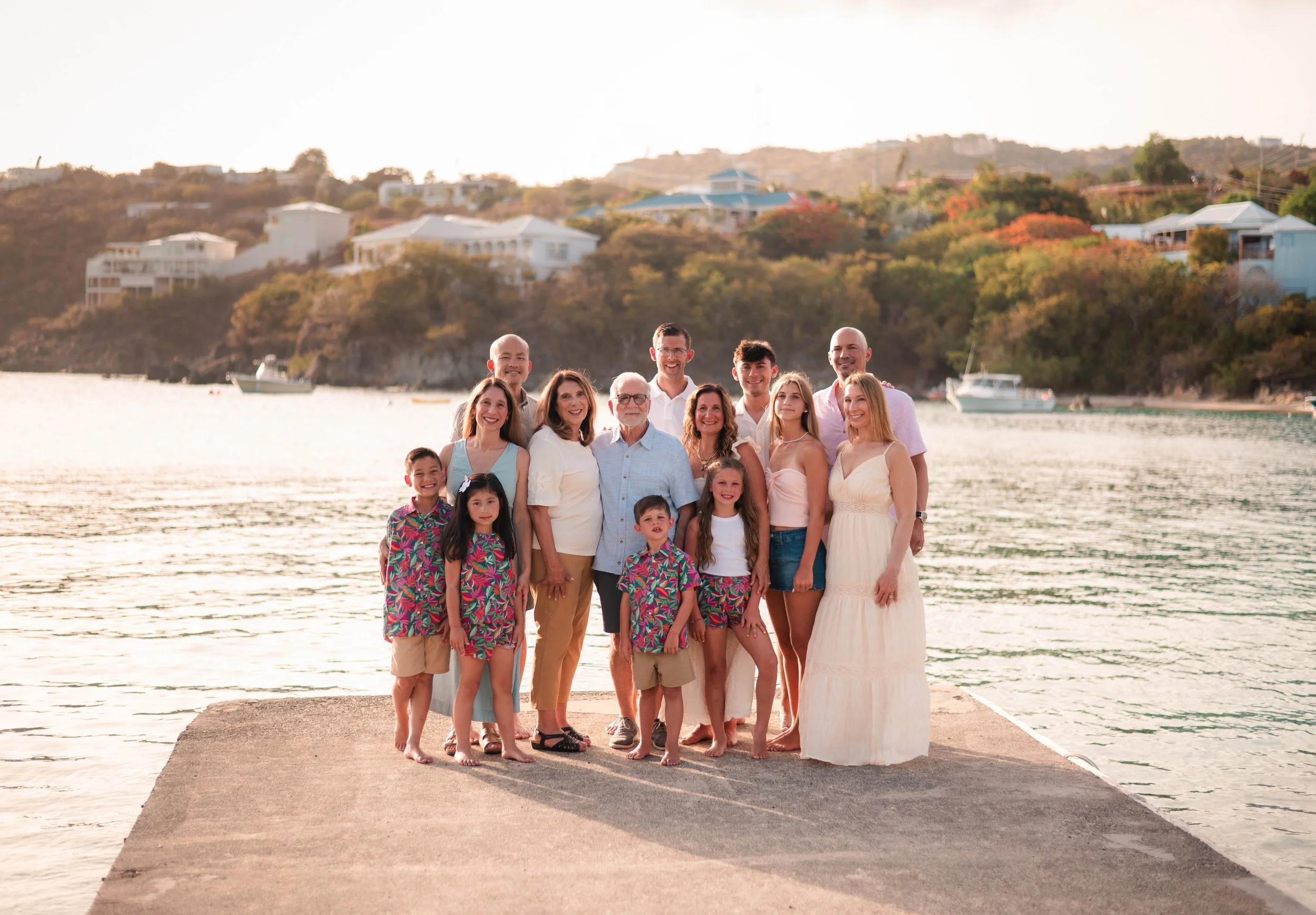 A large family group standing on a concrete pier by the water, smiling, with boats and houses on a hillside in the background during sunset.