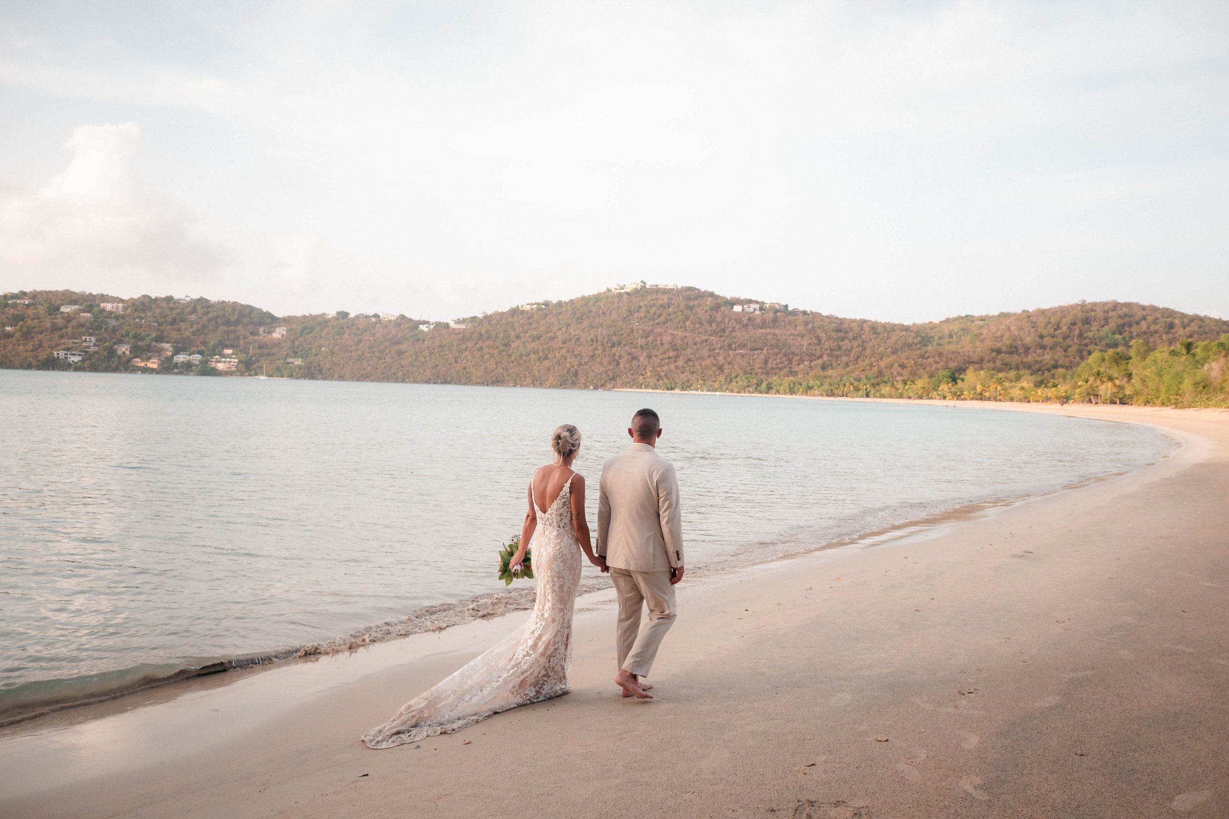 A bride and groom walking hand in hand along a beach shoreline with hills and houses in the background.