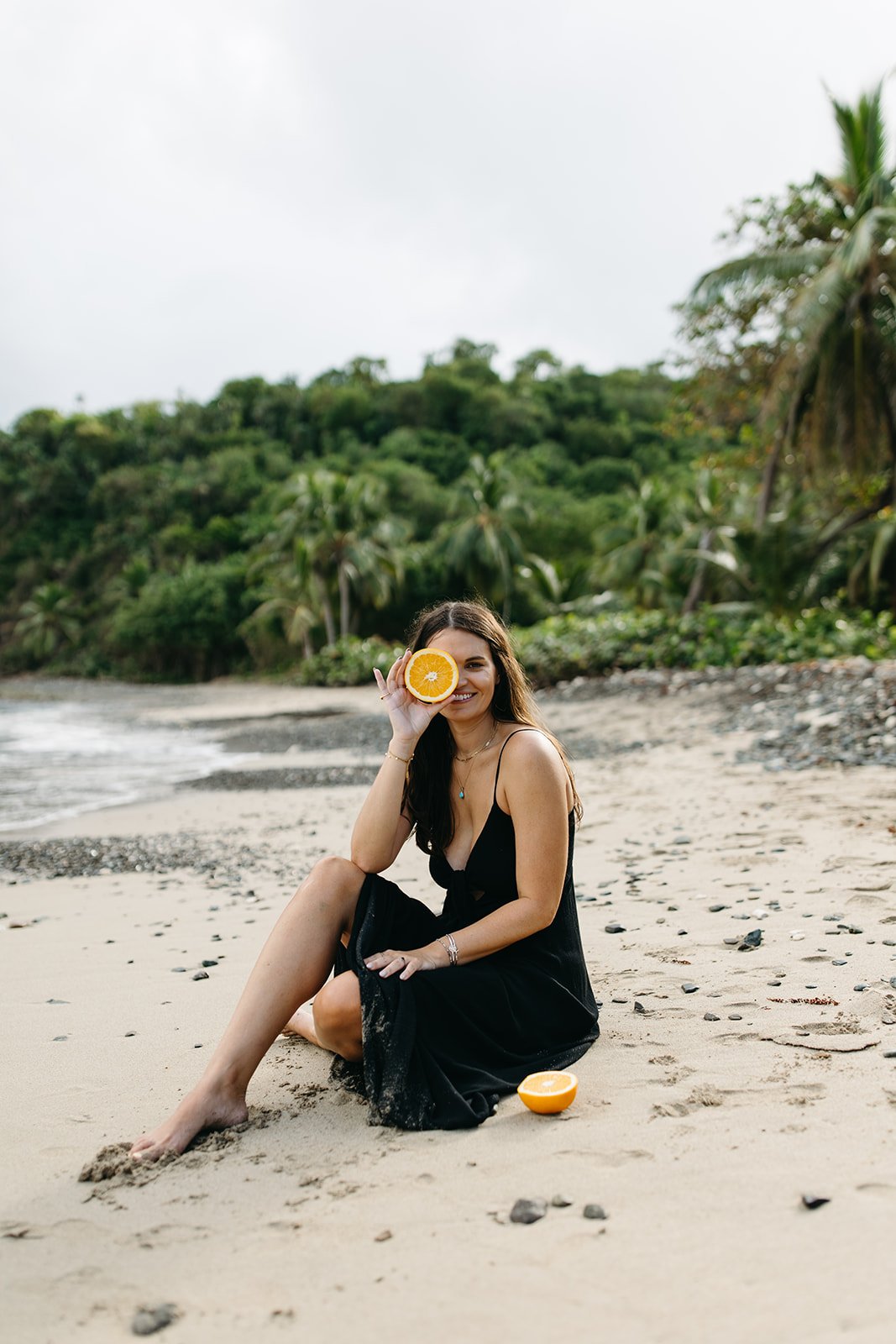 A woman in a black dress sitting on the beach, holding half an orange up to her eye, smiling, with another half orange on the sand beside her, lush greenery and palm trees in the background.