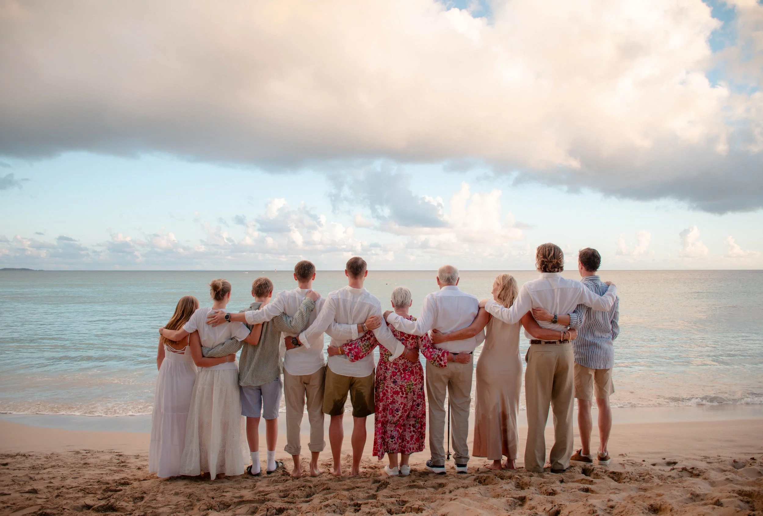 Group of people standing arm in arm on a beach, facing the ocean with cloudy sky overhead.