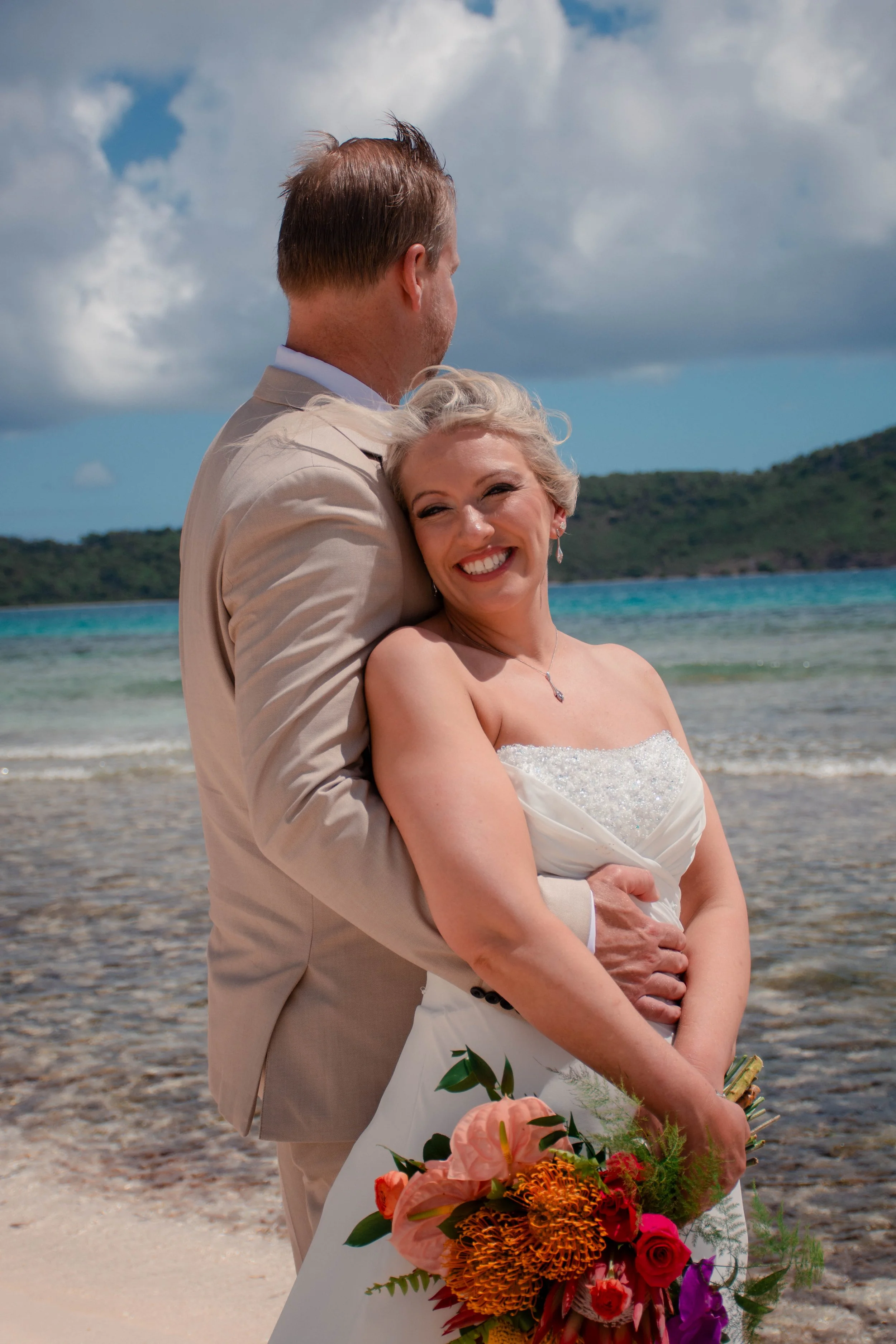 A bride and groom embracing on a beach, smiling with the ocean and hills in the background, she holding a colorful bouquet of flowers.