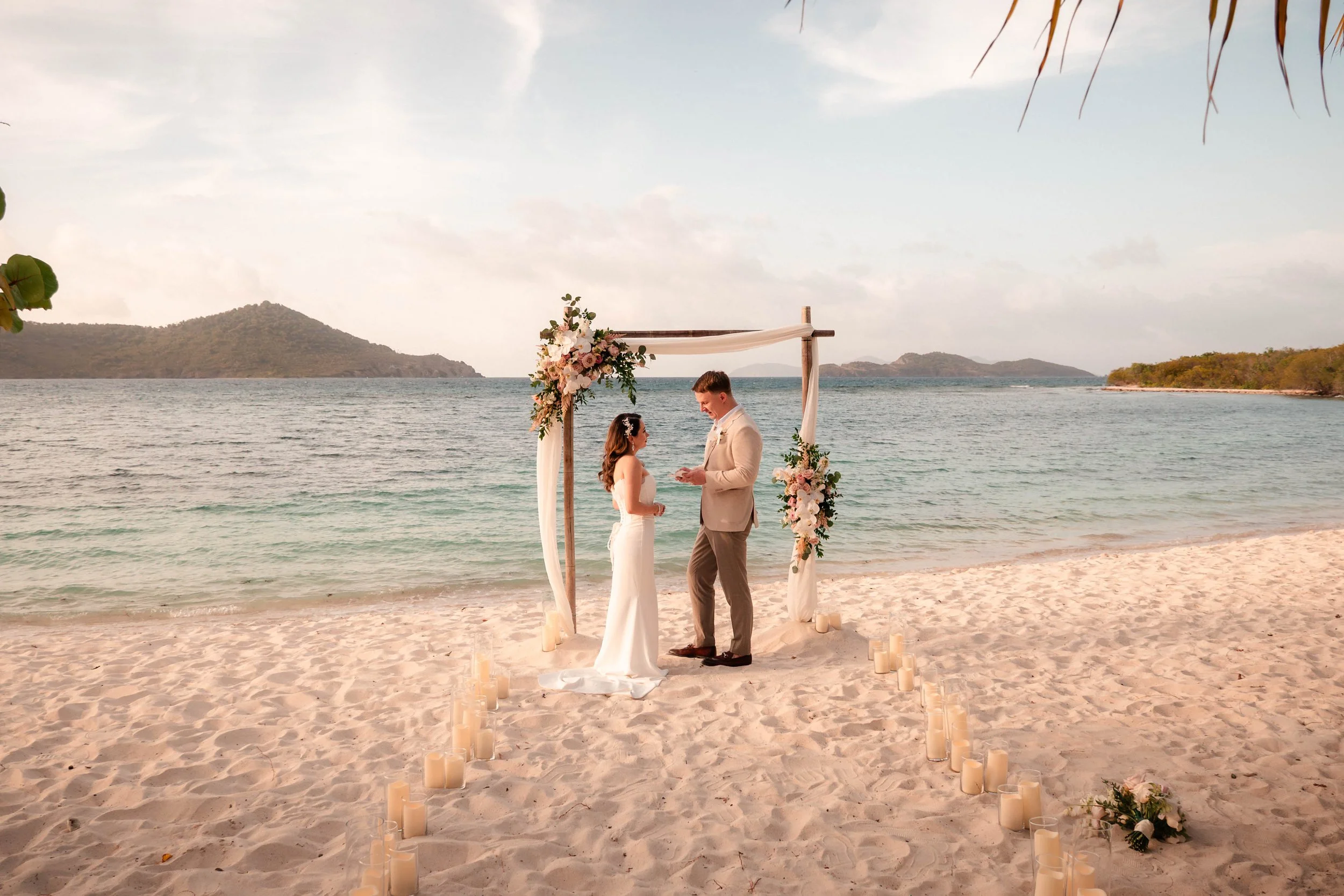 A couple getting married on a beach with an arch decorated with flowers. There are candles surrounding the area, and the ocean with small islands in the background.