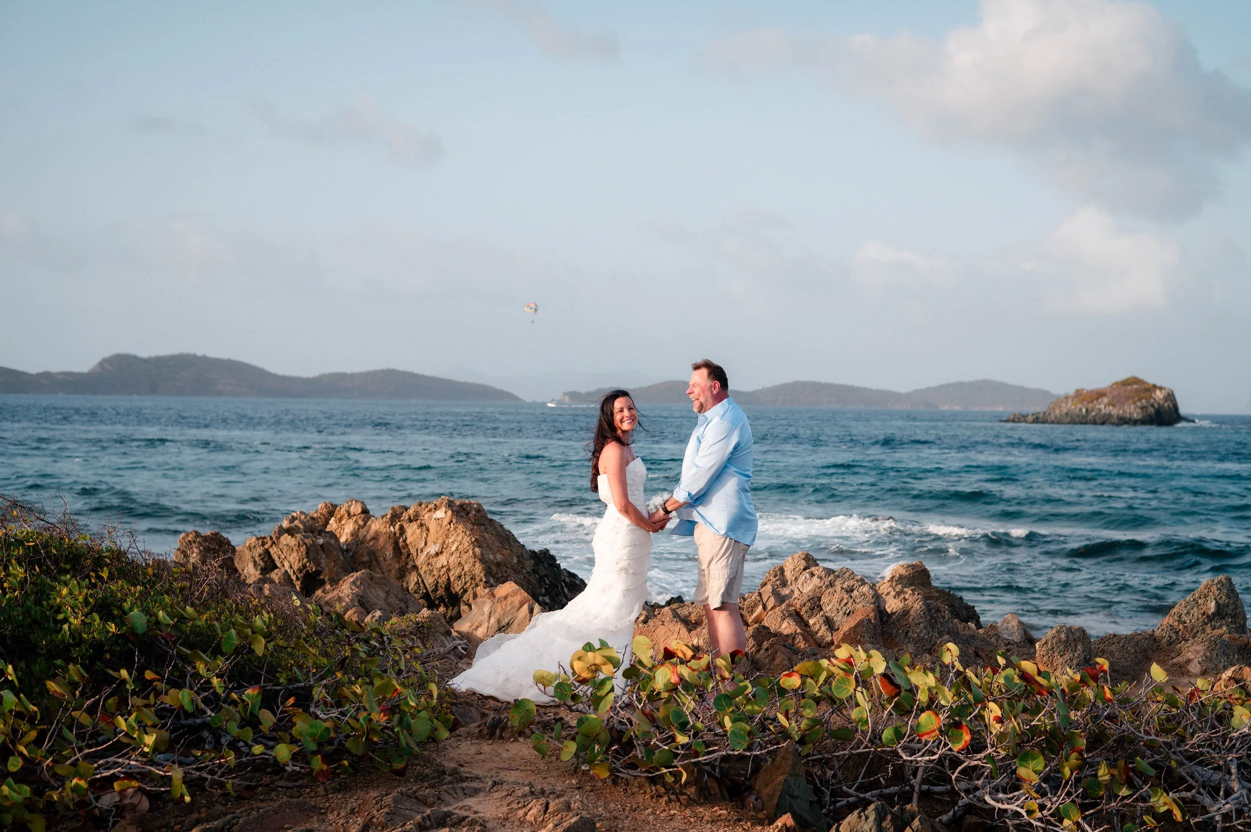 A bride and groom are standing on rocks by the ocean, holding hands and smiling at each other during their wedding. The bride is wearing a white wedding dress, and the groom is dressed in a light blue shirt and beige shorts. There are islands and hil