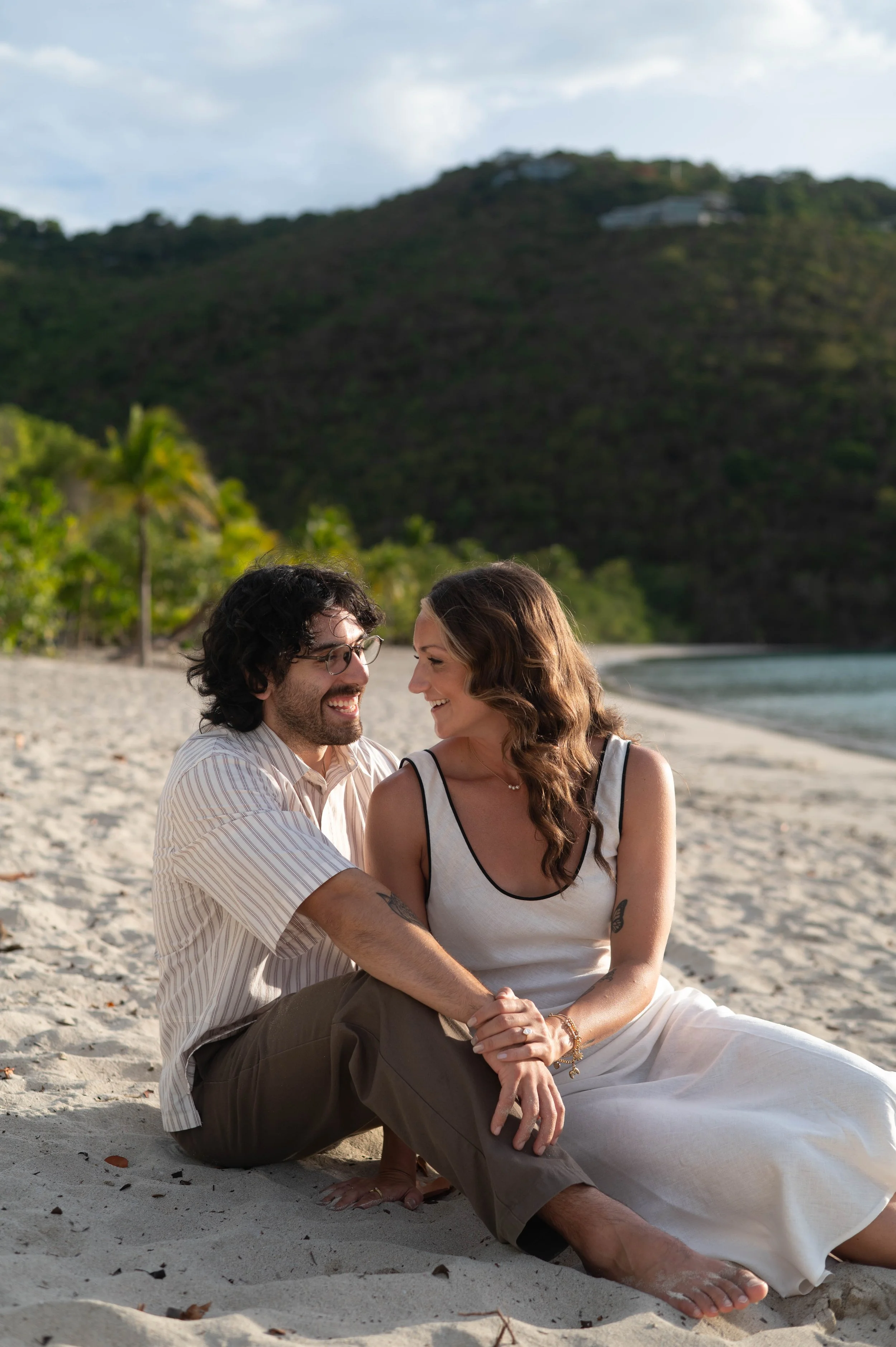 A couple sitting on a sandy beach, smiling and looking at each other, with a hillside and some buildings in the background.