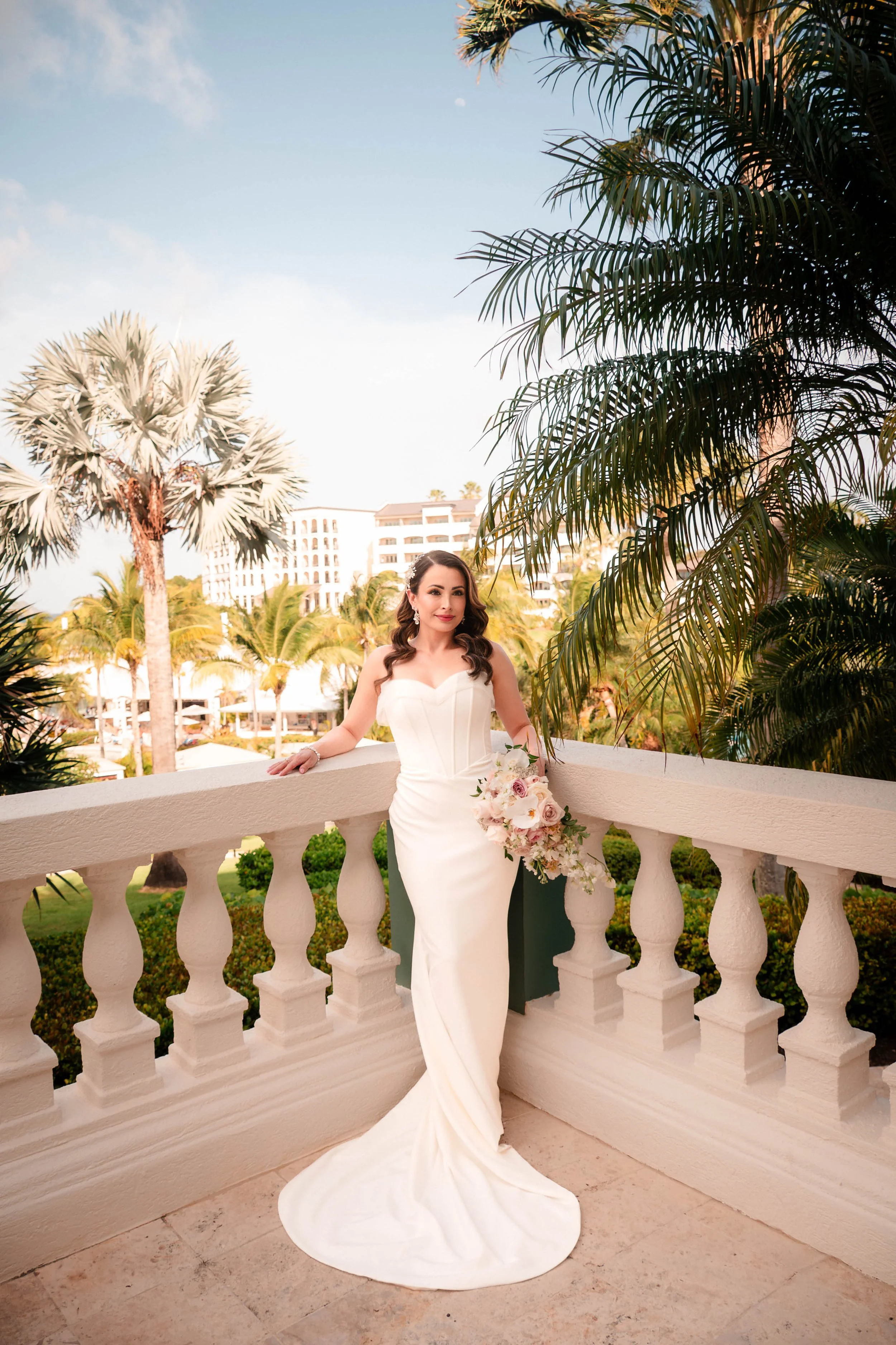 A woman in a white strapless wedding dress holding a bouquet of pale pink and cream roses, standing on a terrace with white balustrade, surrounded by lush tropical greenery and palm trees, with skyscrapers and a clear sky in the background.