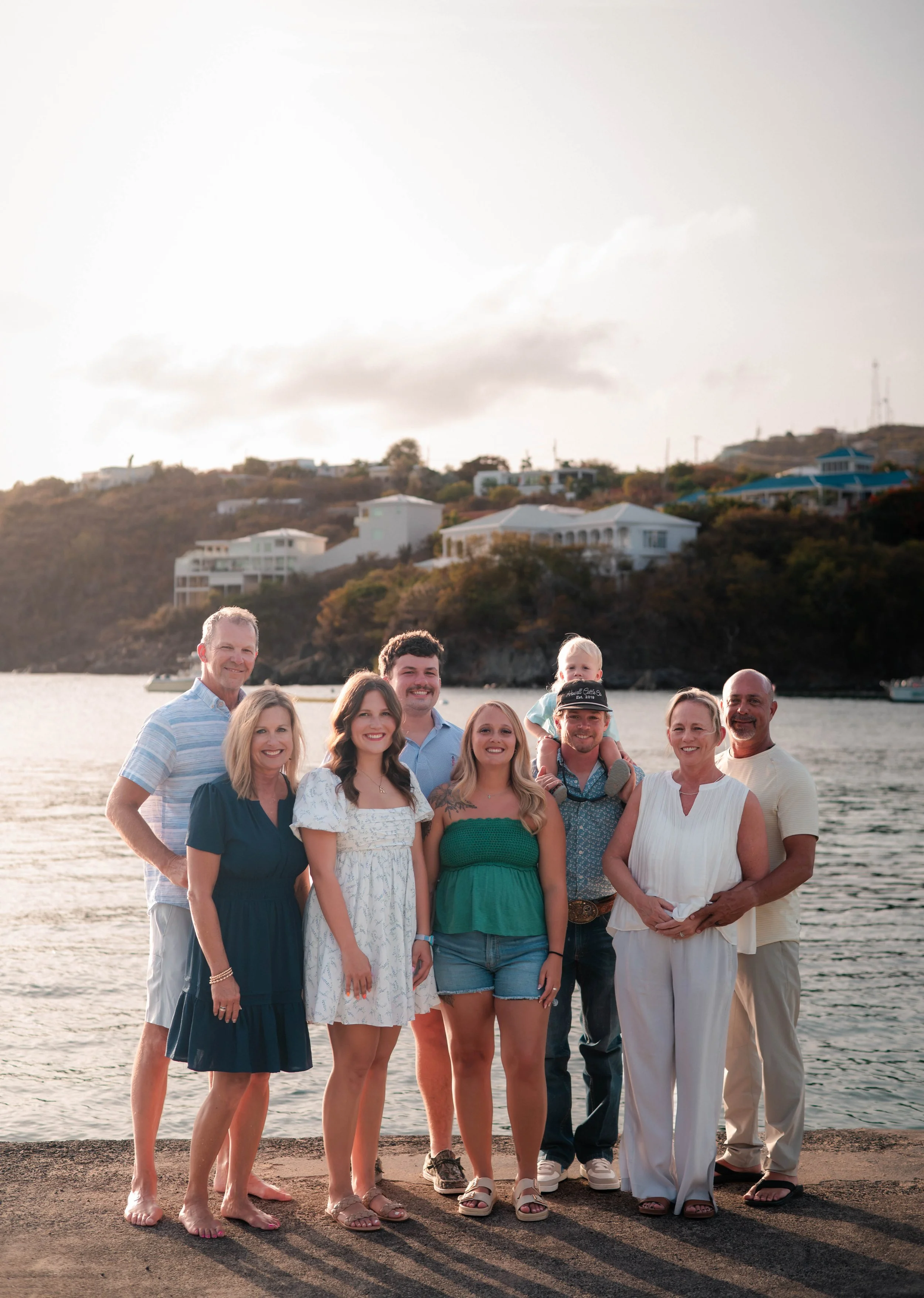 Group of nine people standing on a beach with water and houses on a hill in the background.