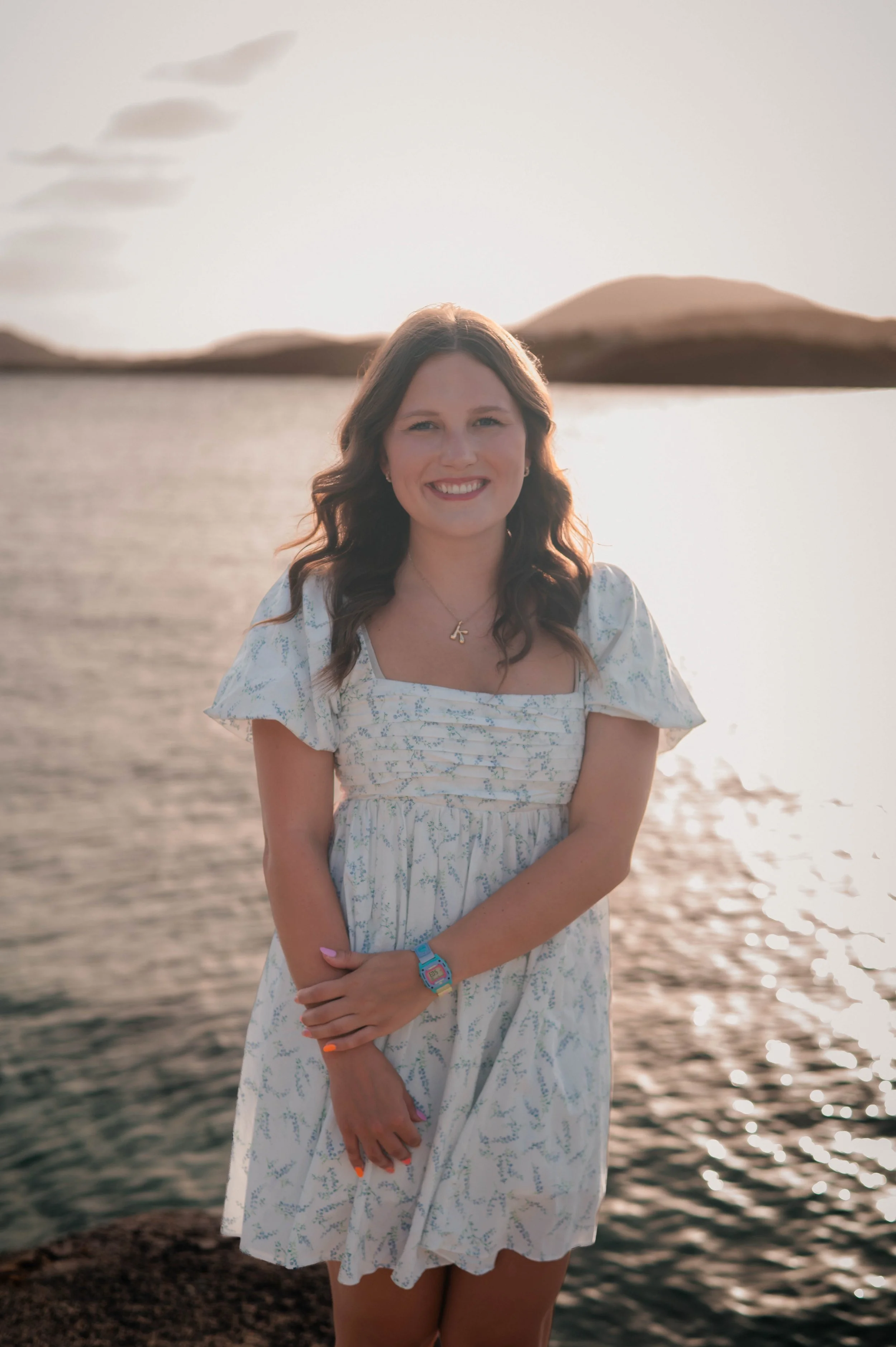 A young woman with brown, wavy hair and a smile standing near a body of water at sunset with hills in the background, wearing a white dress with blue patterns, colorful wristband, and necklace.