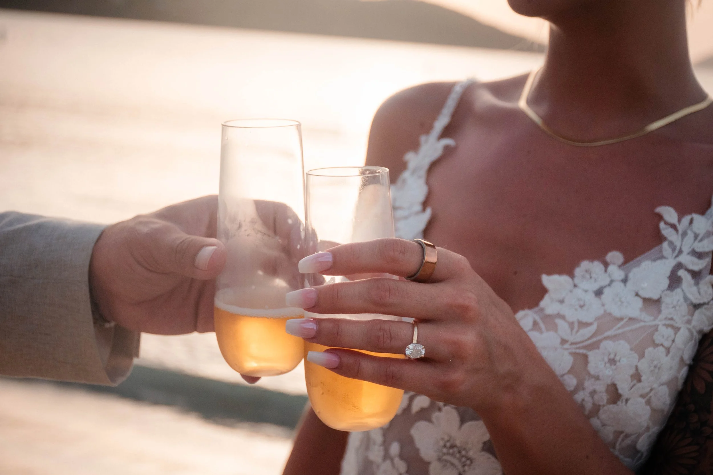 Two women holding glasses of beer and toasting during sunset, one wearing a white lace dress and the other wearing a beige sleeve.