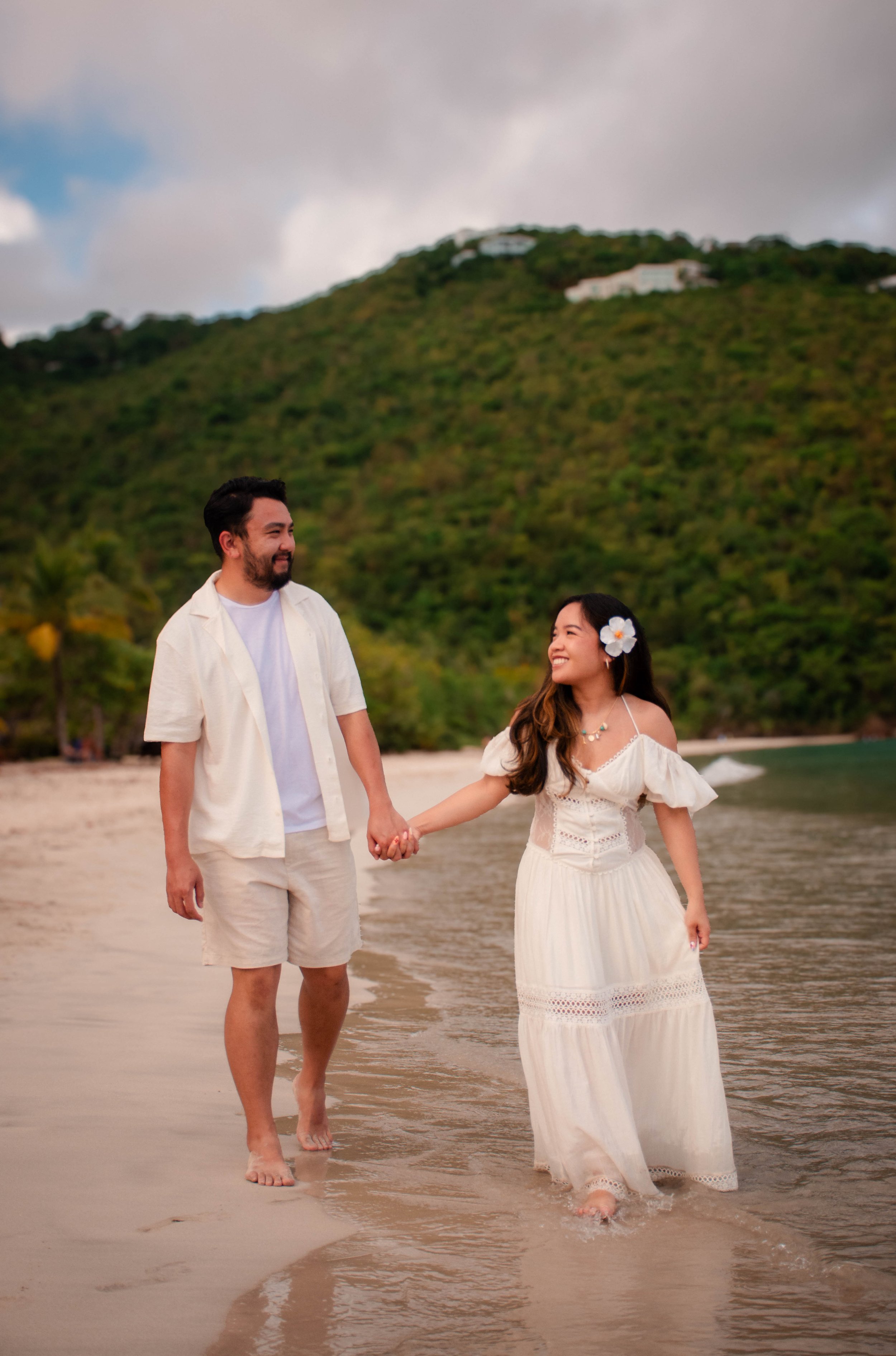 A couple walking hand-in-hand on a beach with green hills in the background, the man wearing a light-colored shirt and shorts, and the woman wearing a white dress with a flower in her hair.