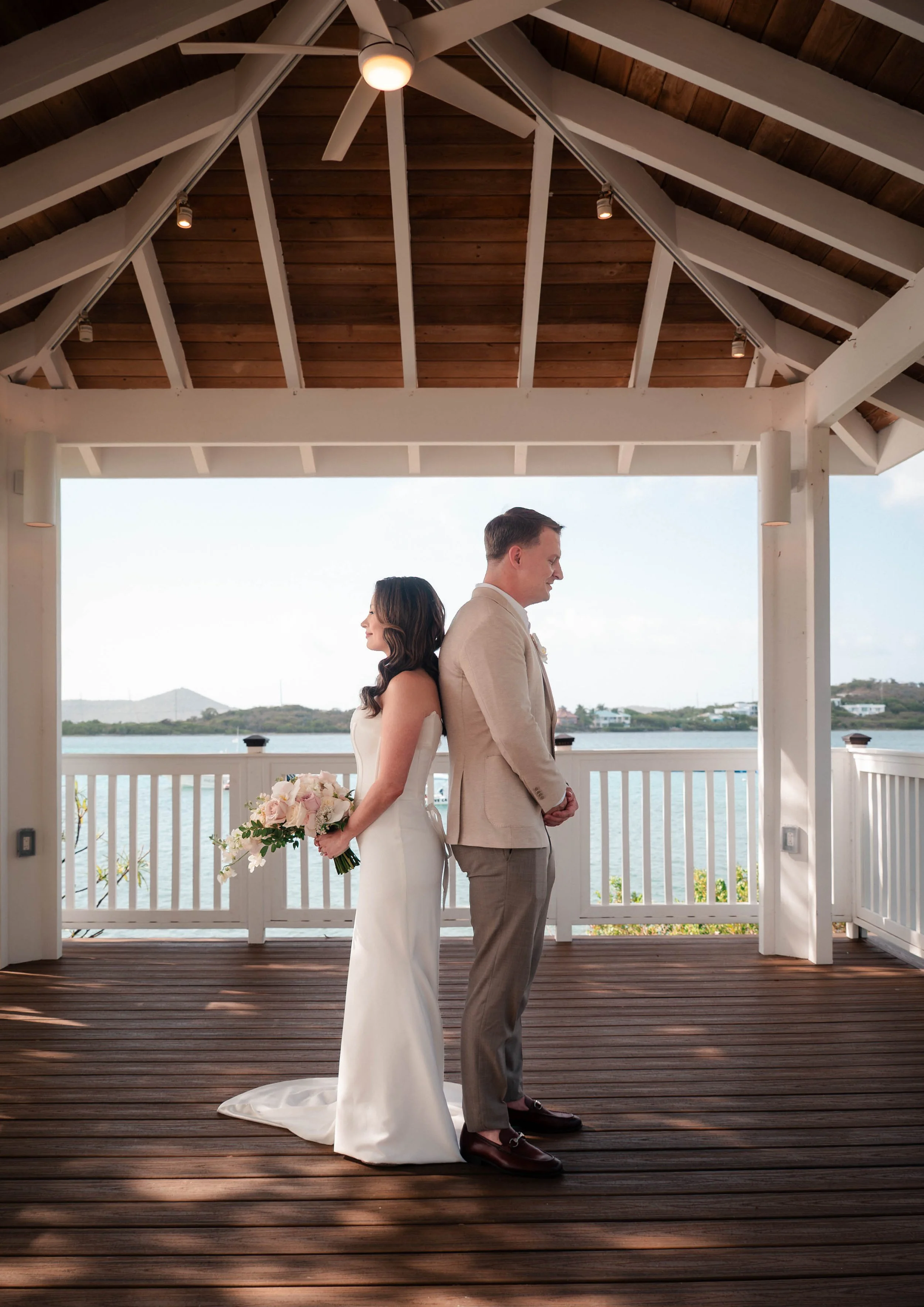 A bride and groom stand back to back on a wooden deck under a covered gazebo with a view of water and hills in the background, during their wedding ceremony. The bride holds a bouquet of pink and white flowers, and the groom wears a beige suit and da
