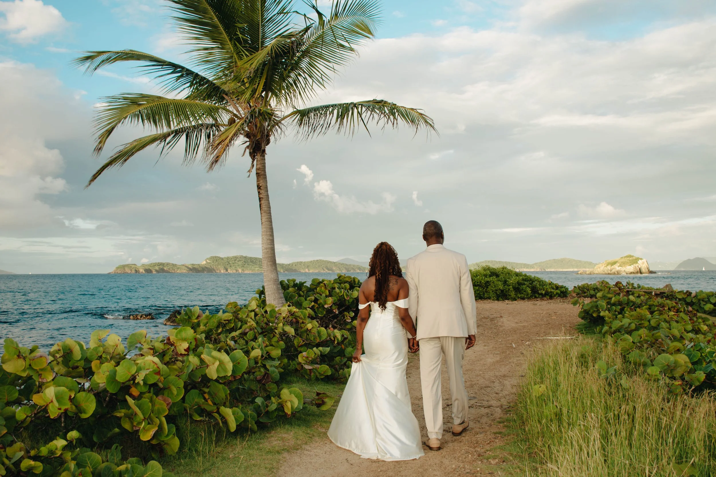 A couple dressed in wedding attire walking by the ocean on a beach, with greenery and a palm tree in the foreground, and distant islands in the background.