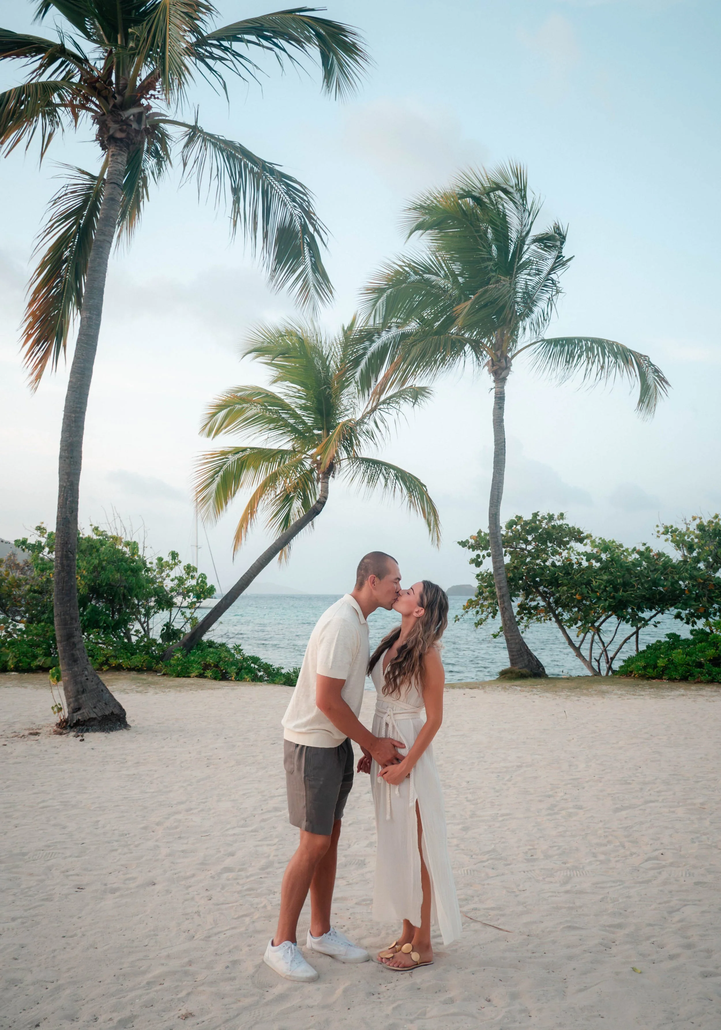 A couple sharing a kiss on a sandy beach with palm trees and an ocean view in the background.