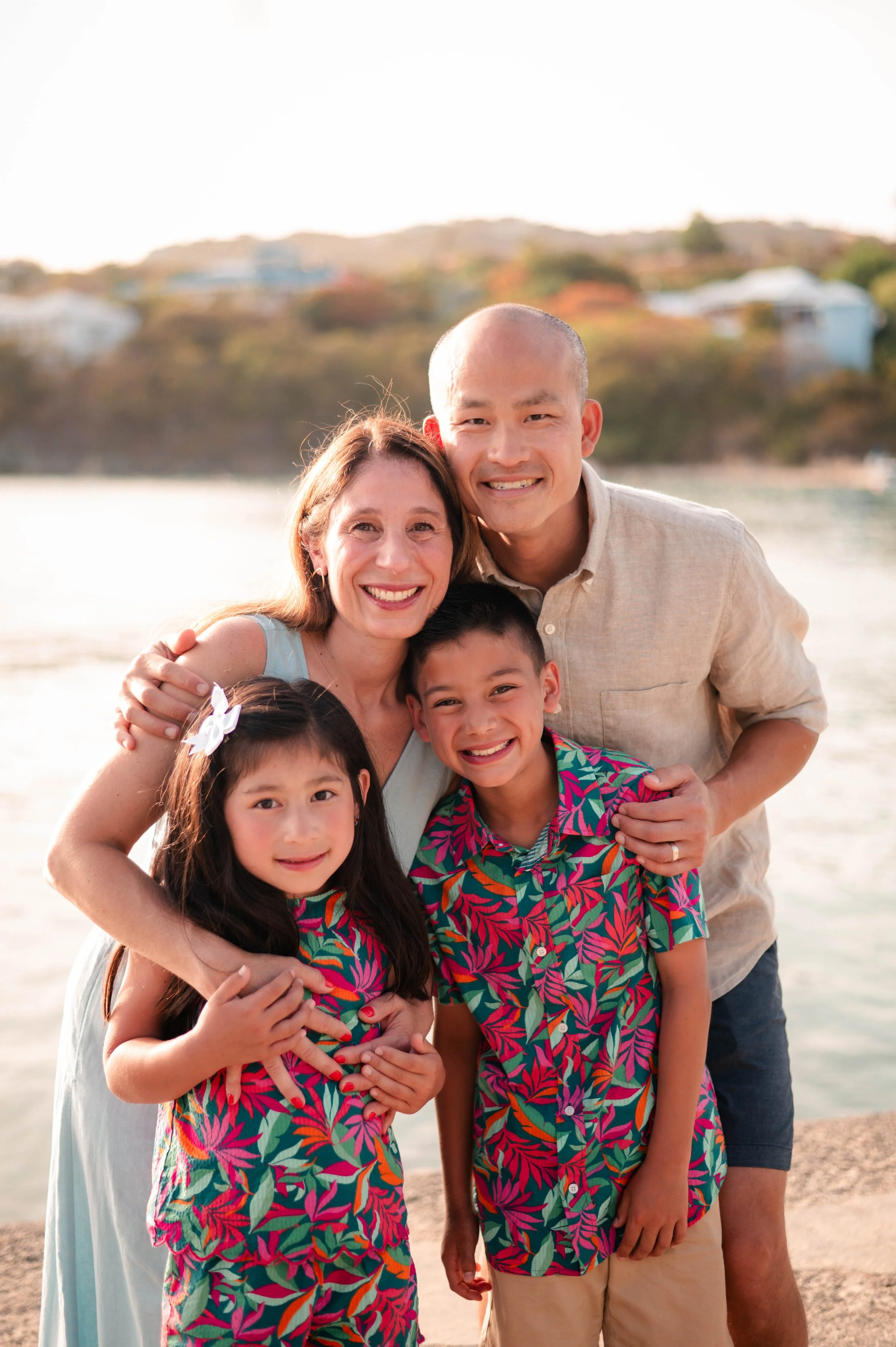 A group of five people, including two adults and three children, smiling and hugging on a beach with a hillside and houses in the background during sunset.