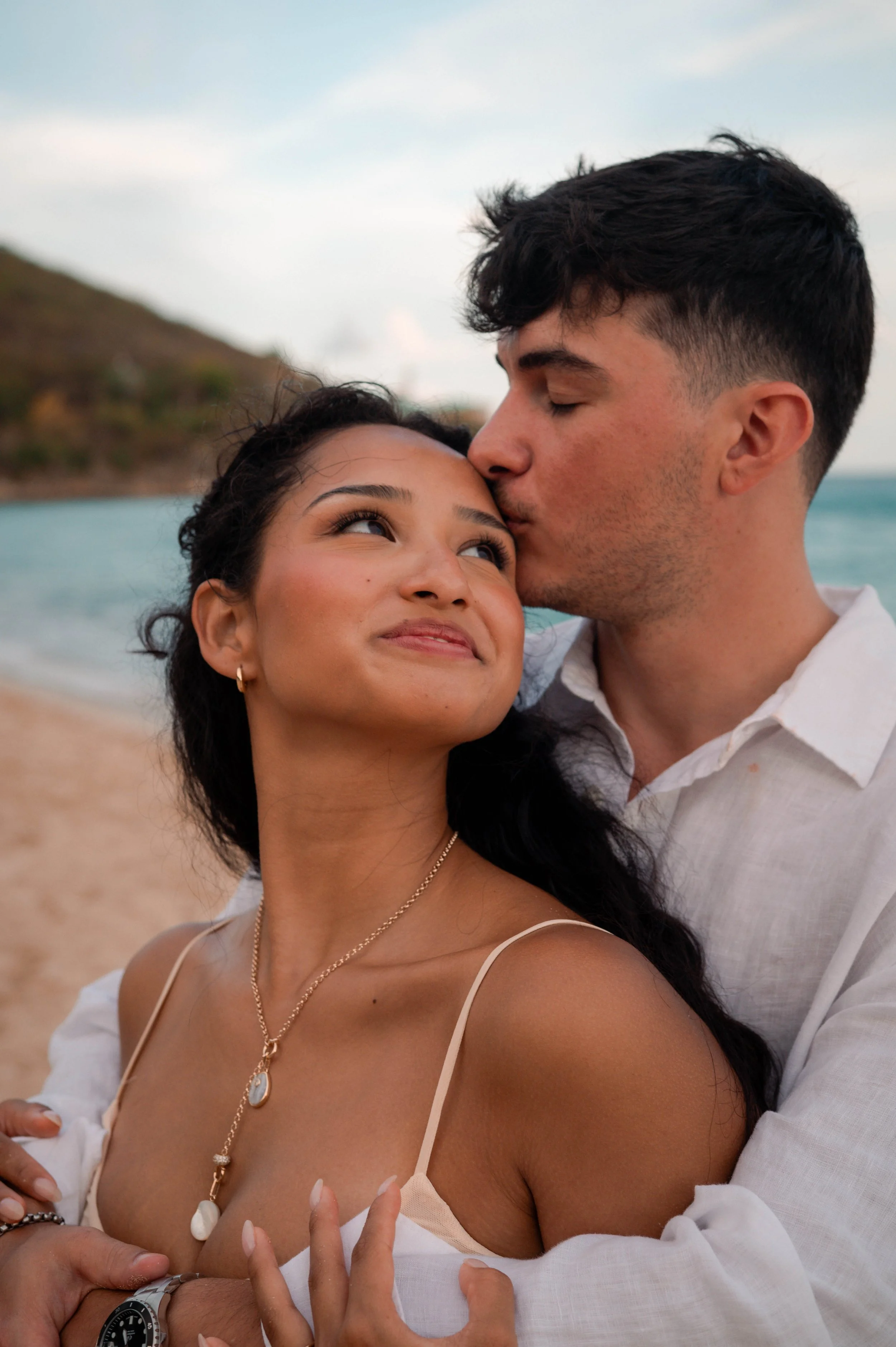 A young woman and a young man are close together on a beach, with the woman looking up and smiling while the man kisses her forehead. The background shows a shoreline with hills and the ocean.