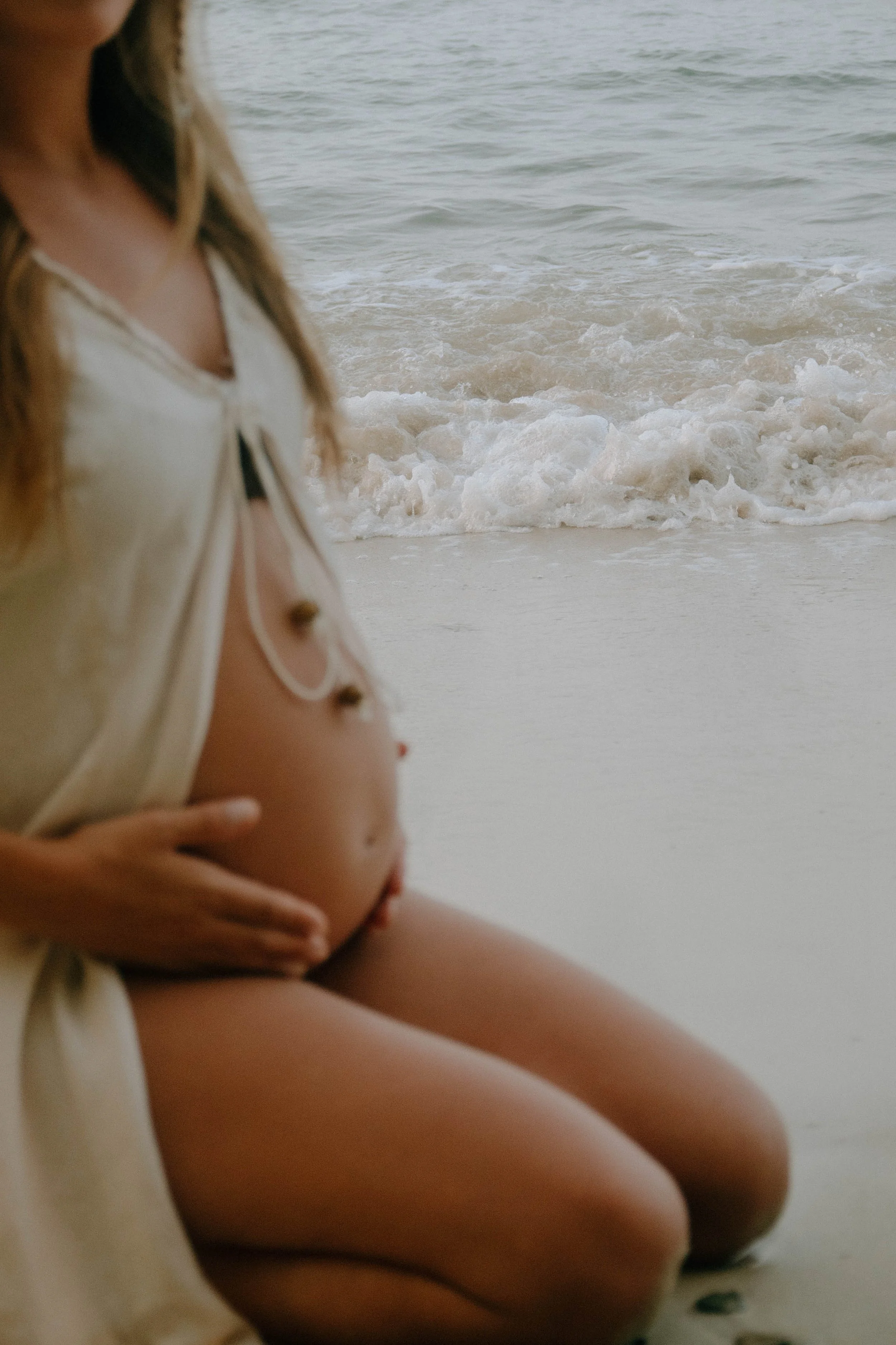A pregnant woman sitting on the beach, with her hand on her belly, wearing a white, loose shirt, and looking away from the ocean.