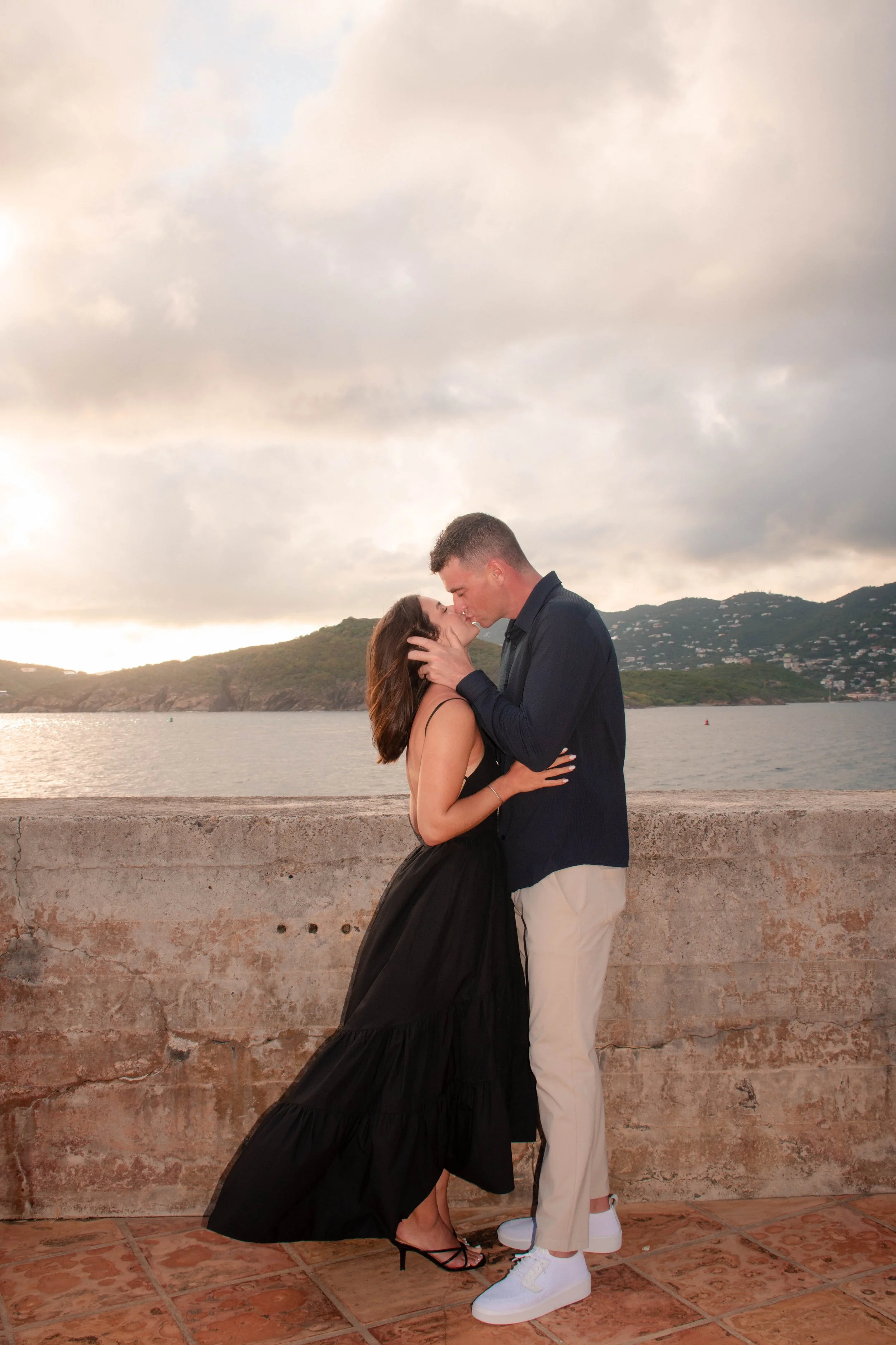 A couple is kissing by a waterfront with hills and houses in the background, during cloudy sunset.