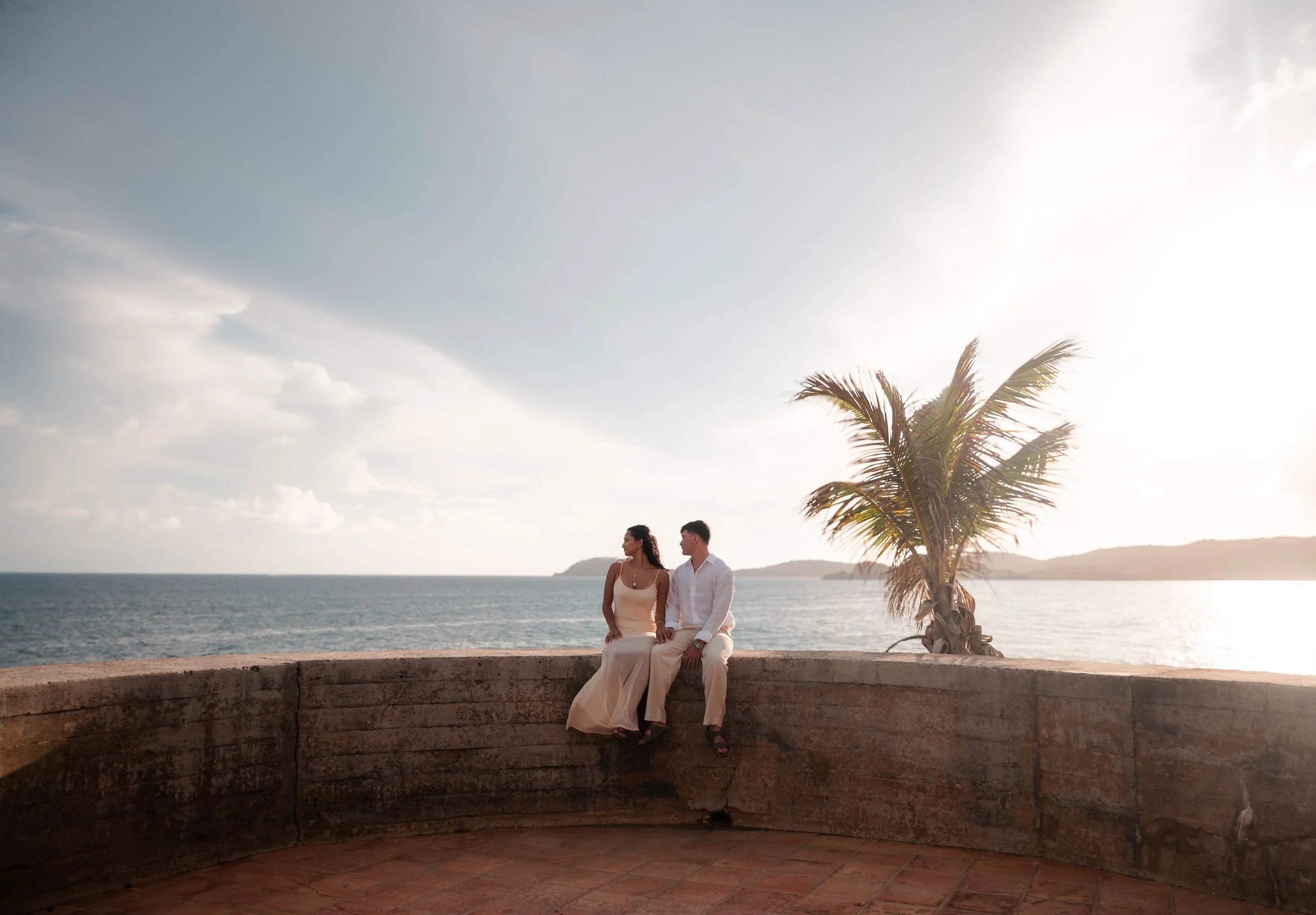 A couple sits on a stone wall by the water, with a palm tree on the right and a cloudy sky, near sunset.
