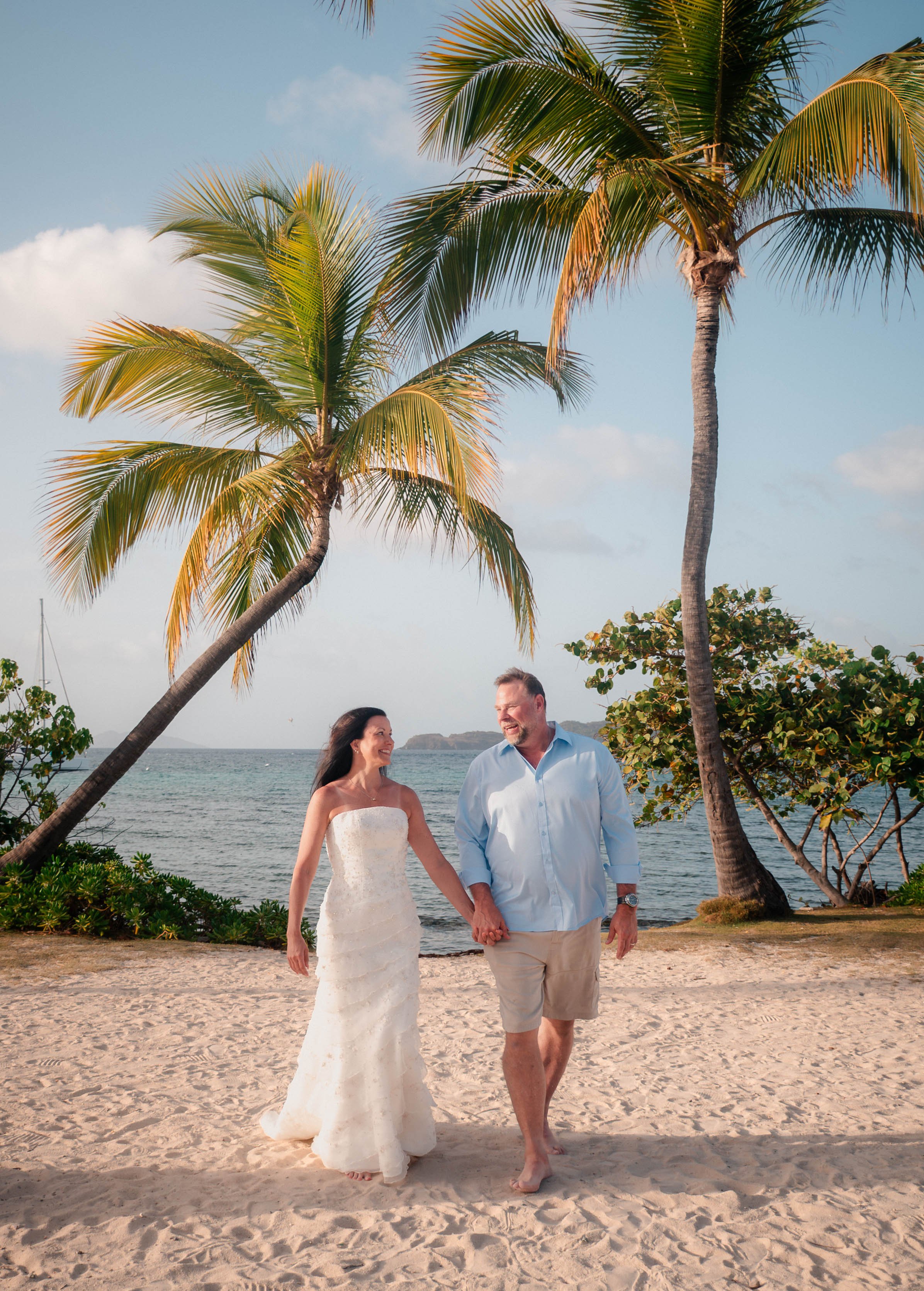 A couple walking hand in hand on a beach with palm trees, ocean, and blue sky in the background, dressed in wedding attire.