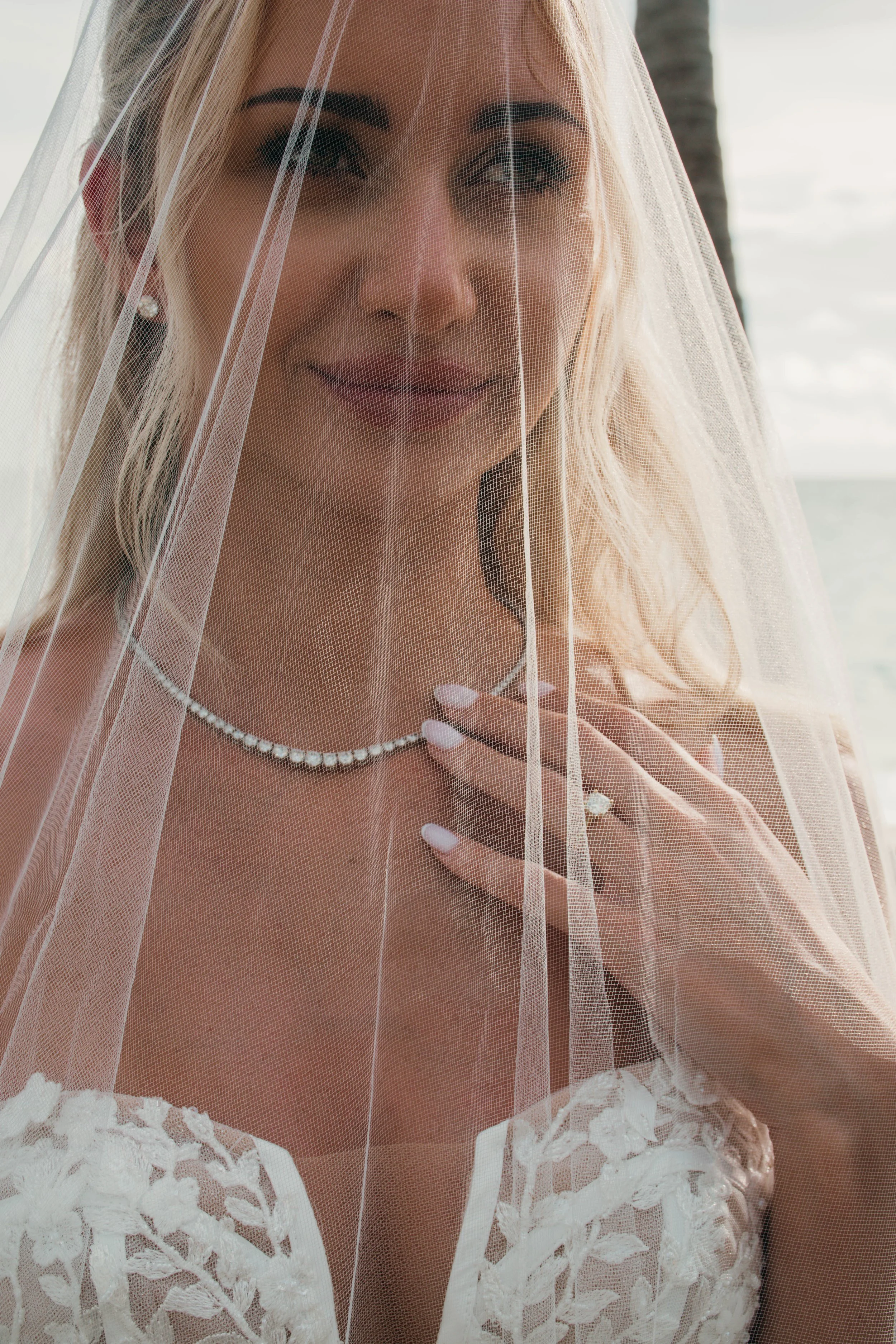 A woman in a wedding dress with lace details, wearing a pearl necklace and earrings, partially covered by a sheer veil, standing outdoors near a body of water with a palm tree in the background.
