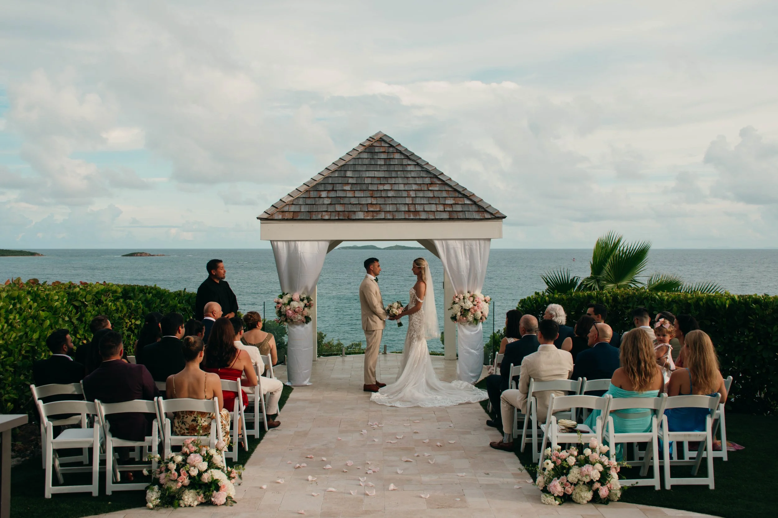 A wedding ceremony taking place outdoors by the ocean, with a groom and bride standing under a gazebo, surrounded by guests seated in white chairs, with floral decorations and an ocean view in the background.