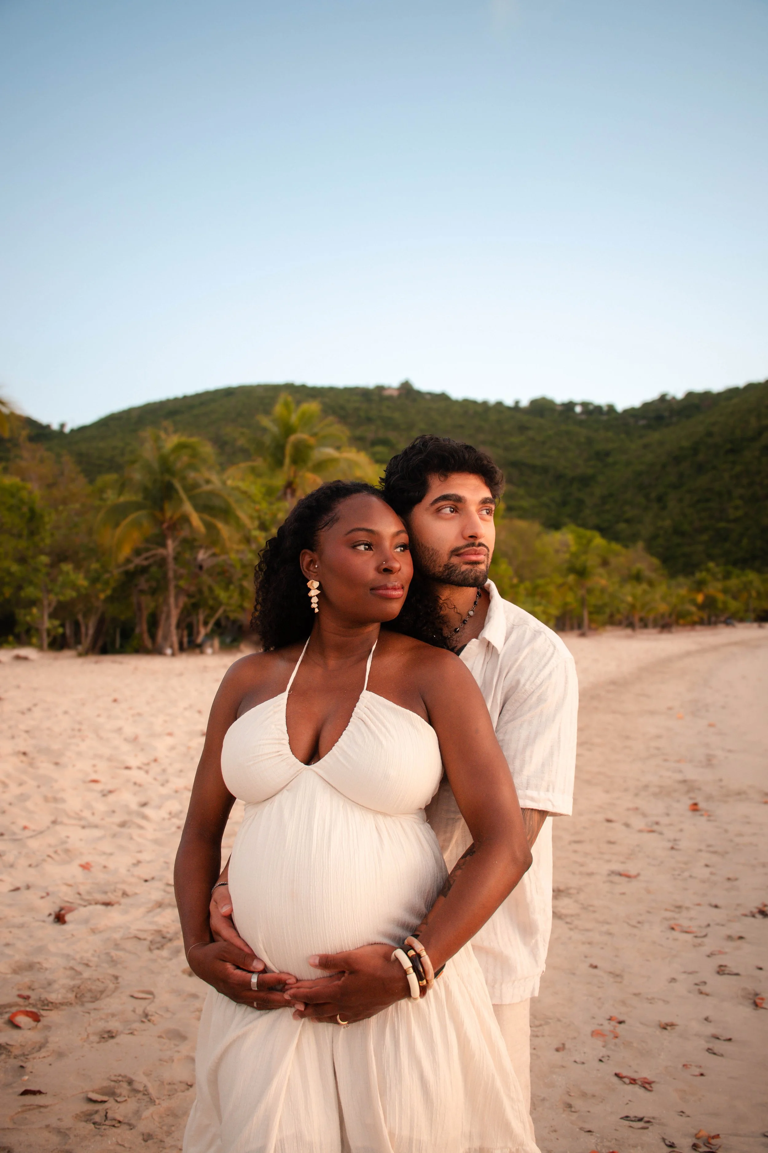 A couple, a pregnant woman and a man, standing on a beach with a lush green hillside in the background during sunset, embracing each other.