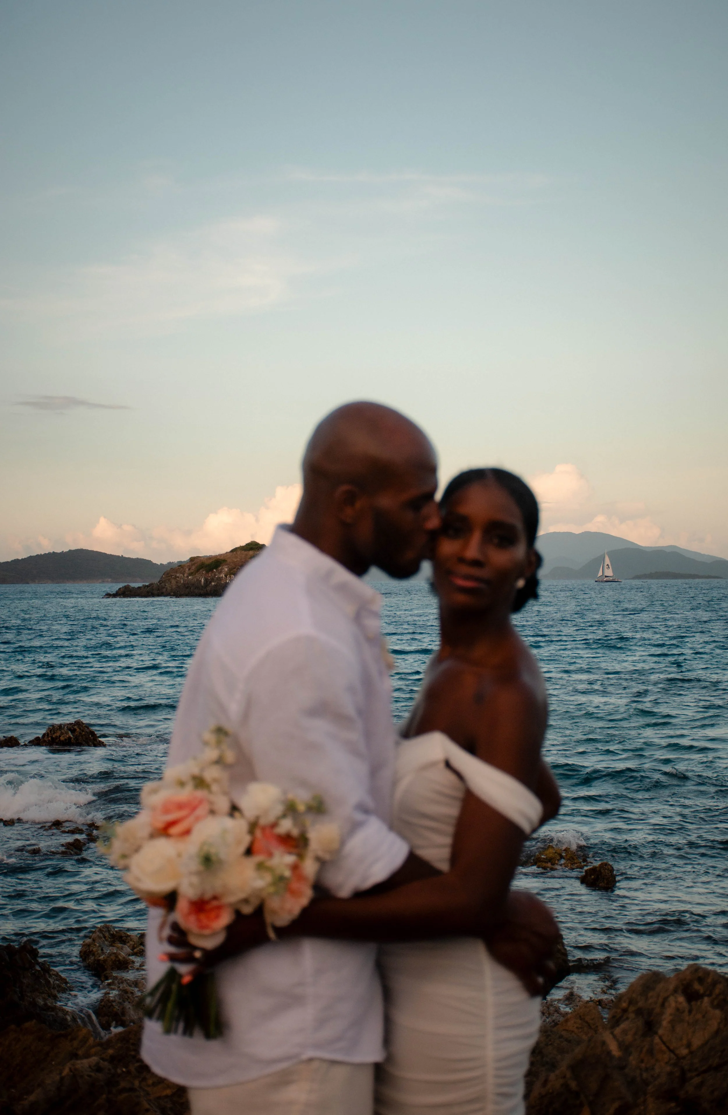 A couple, dressed in white, stands close together near the ocean during sunset, with mountains and a sailboat in the background. The man holds a bouquet of white and pink flowers.