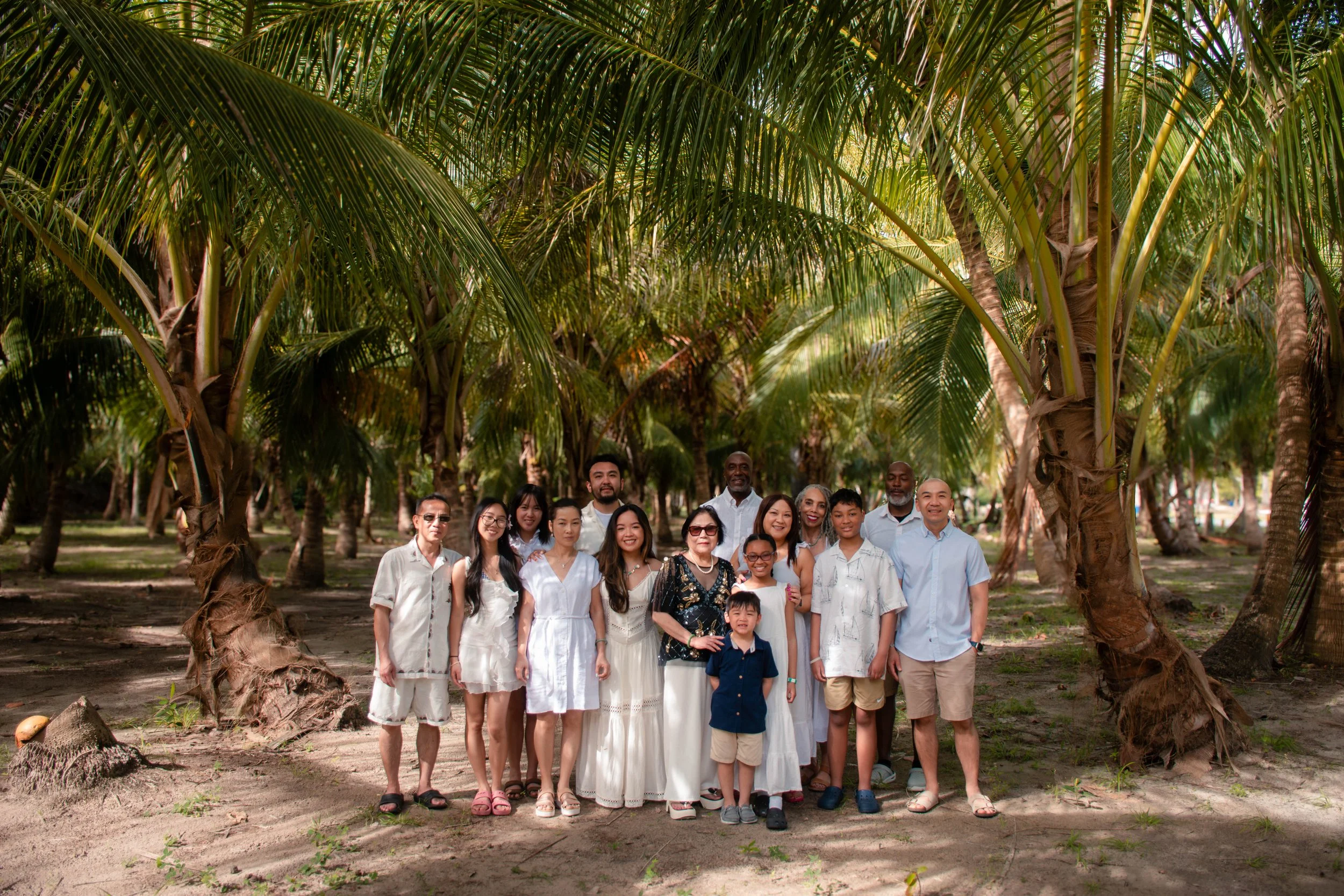 A large group of diverse people, including men, women, and children, standing in a line outdoors under tall palm trees, with greenery and sunshine in the background.