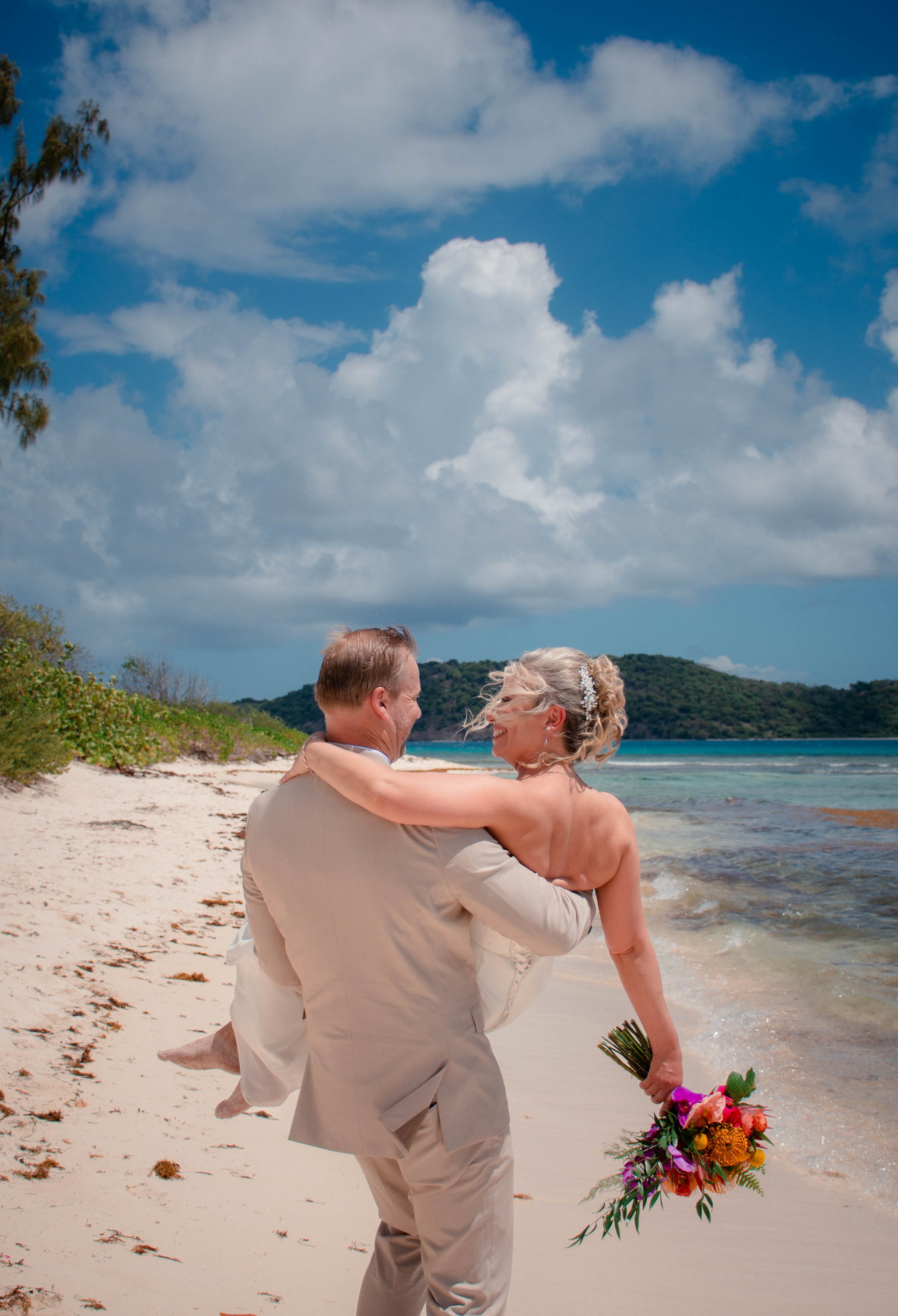 An older couple embracing and smiling on a beach with the ocean in the background, one holding a bouquet of colorful flowers.