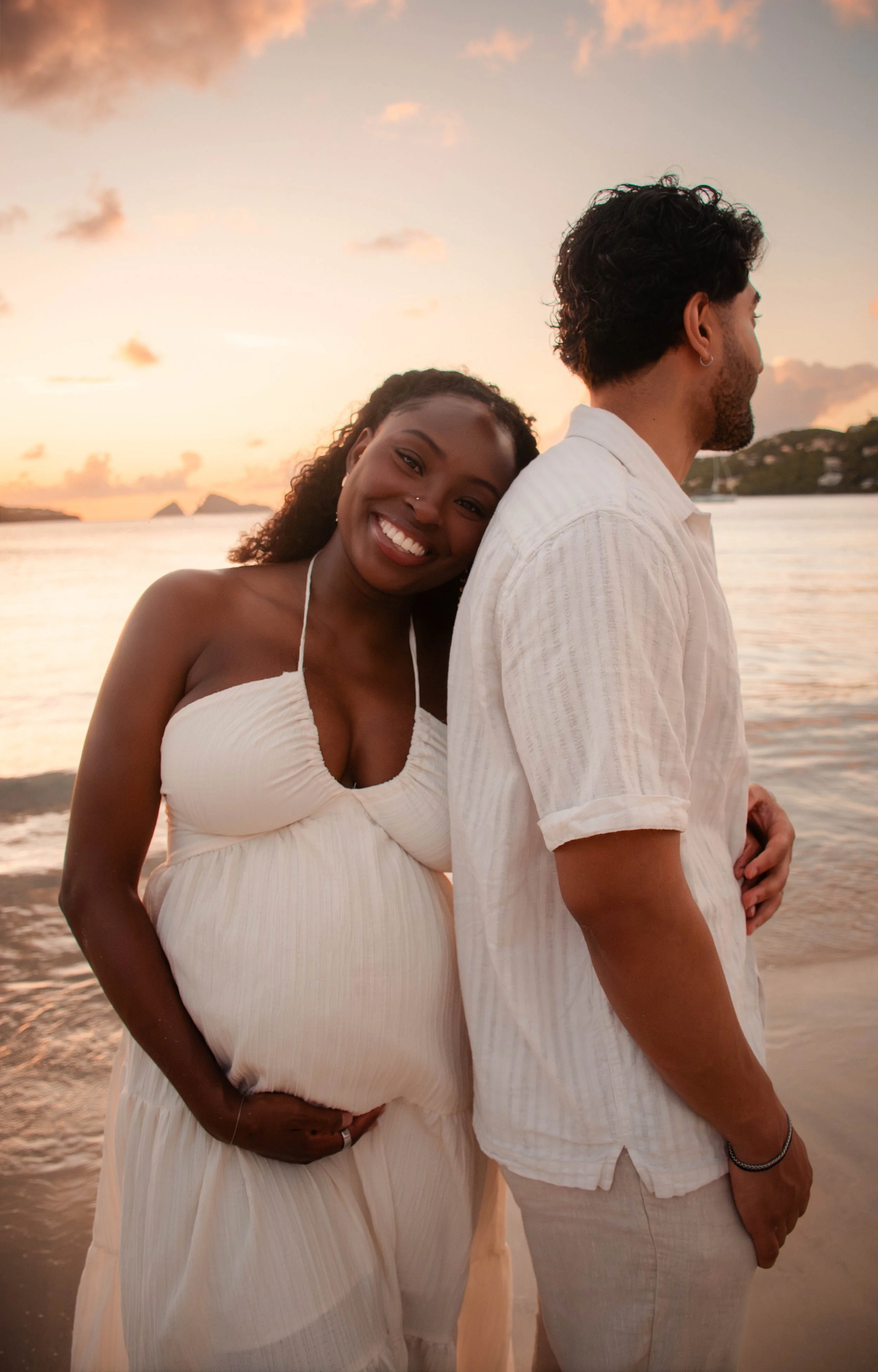 A smiling woman in a white dress hugging a man from behind on a beach at sunset, with water and islands in the background.
