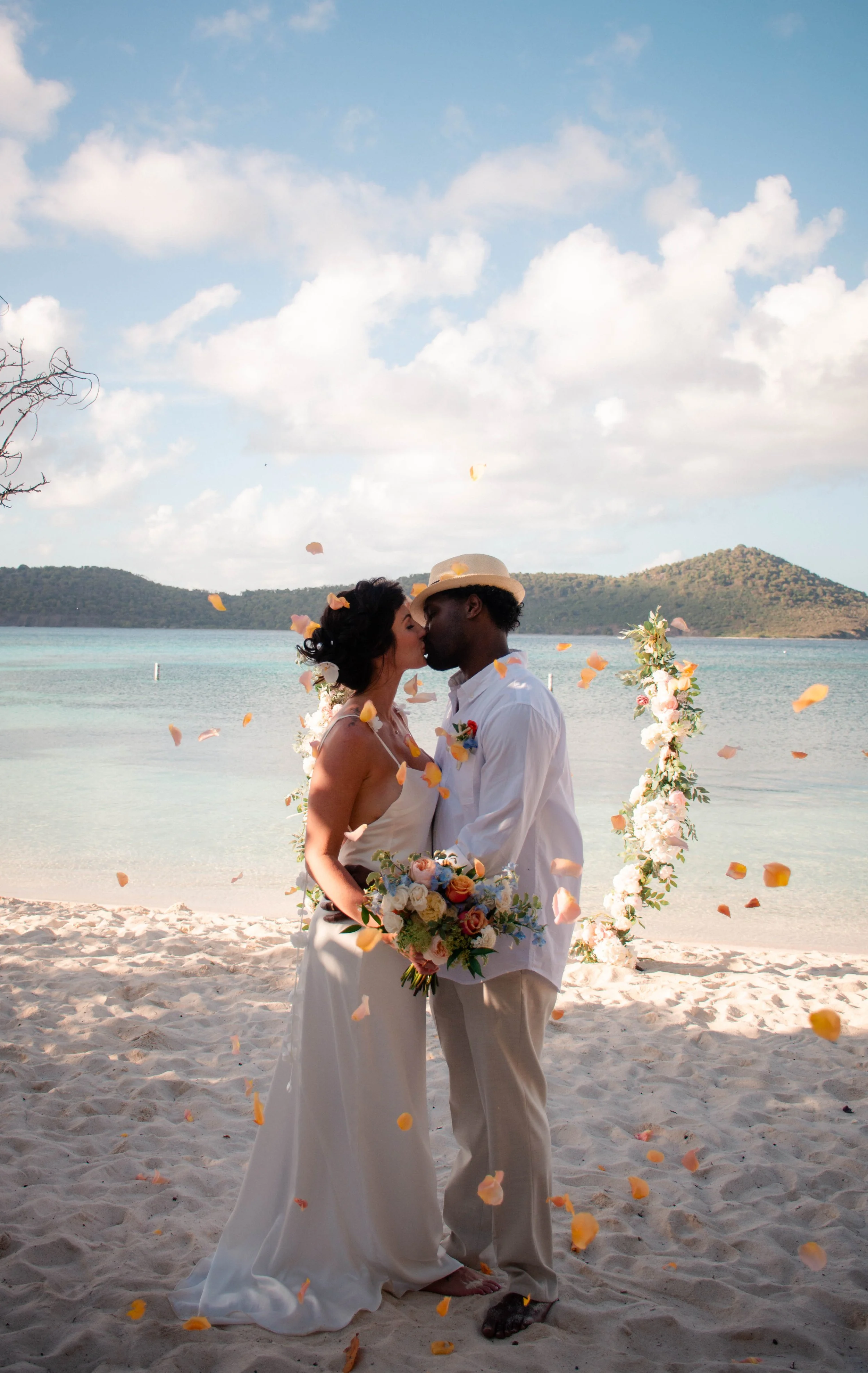 A couple in wedding attire sharing a kiss on a beach, with a floral wedding arch and falling flower petals, ocean, and hills in the background.