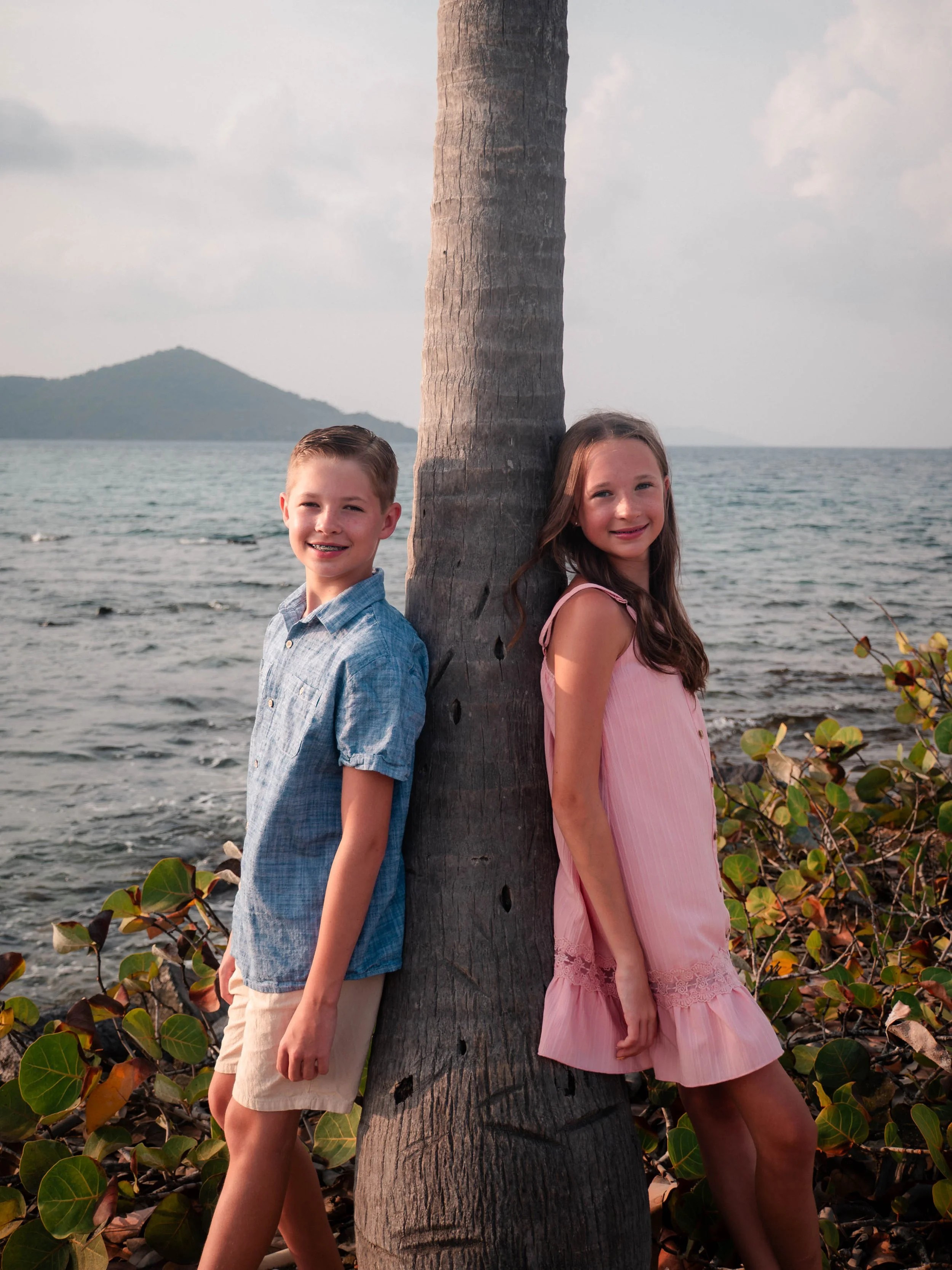 A boy and girl are standing back to back on either side of a tree trunk at the beach. The boy is smiling and wearing a blue checked shirt, while the girl is also smiling, wearing a pink dress. The ocean and a landmass are visible in the background.