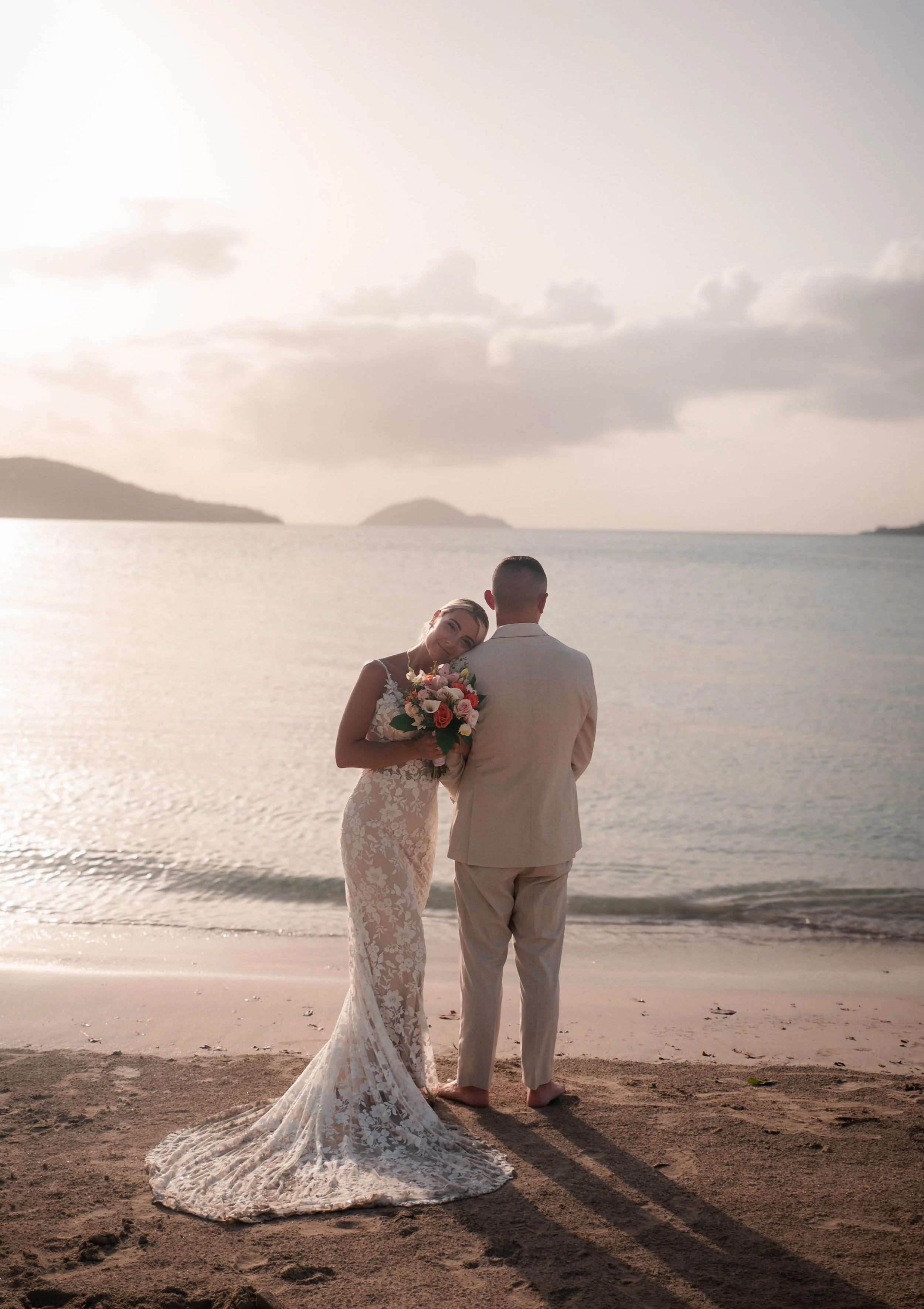 A bride and groom standing on a beach at sunset, with the bride resting her head on the groom's shoulder and holding a flower bouquet, both facing the water.