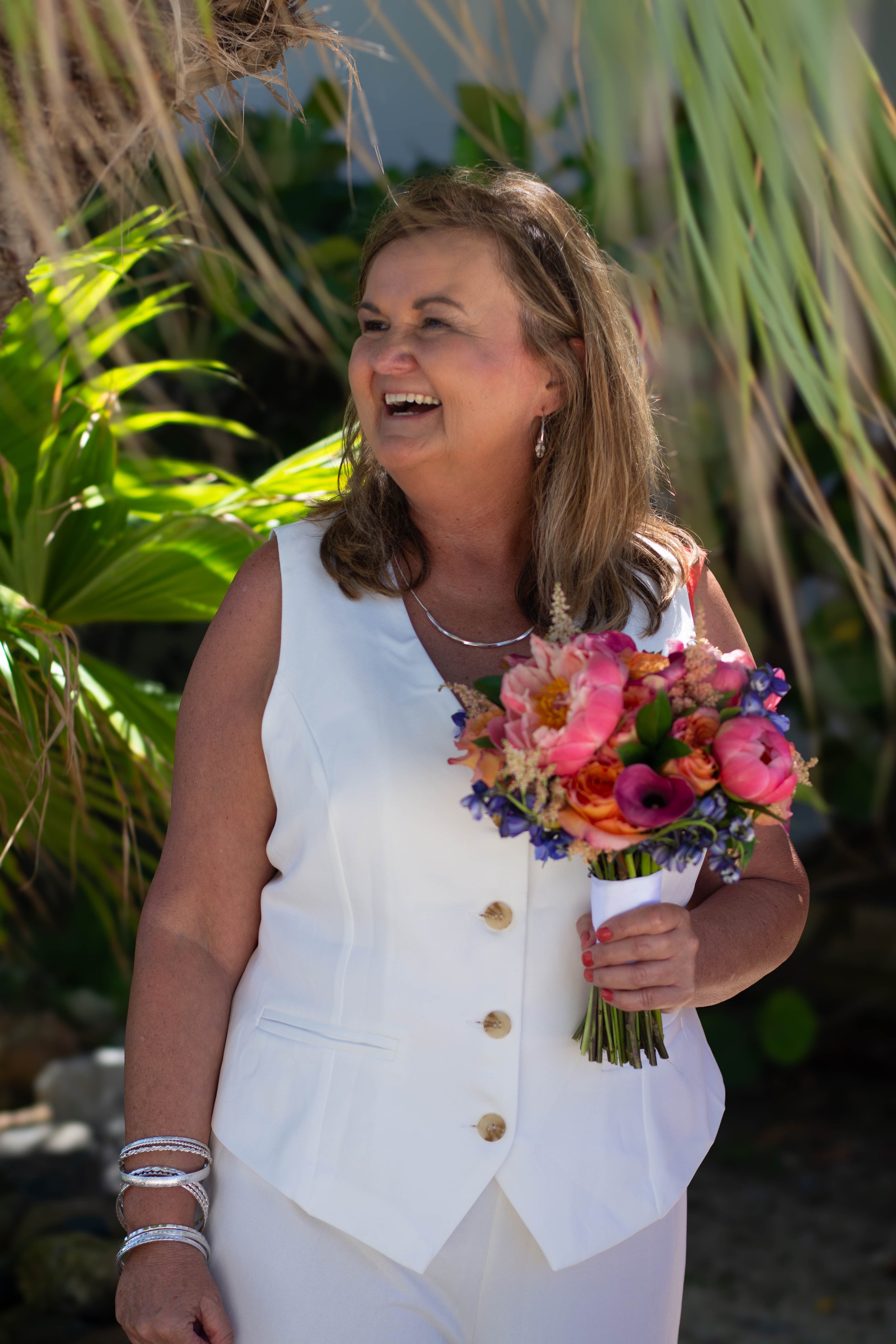 A woman in a white sleeveless dress with large buttons, holding a colorful bouquet of flowers. She is smiling and standing outdoors among green tropical plants.