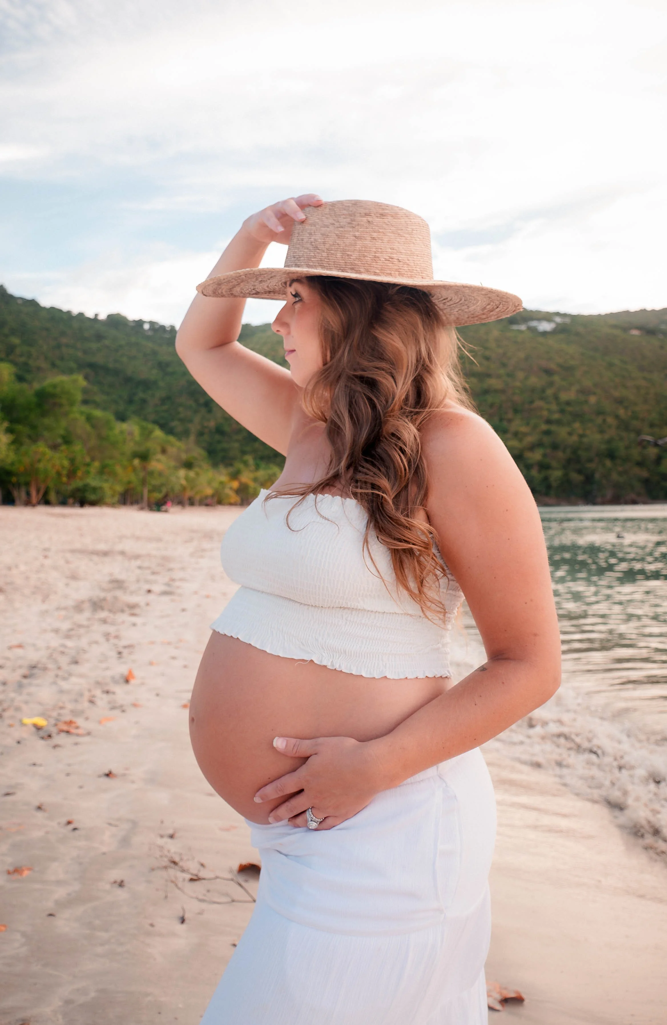 Pregnant woman with long brown hair wearing a straw hat and white dress on a beach, holding her belly with one hand, with green hills and blue sky in background.