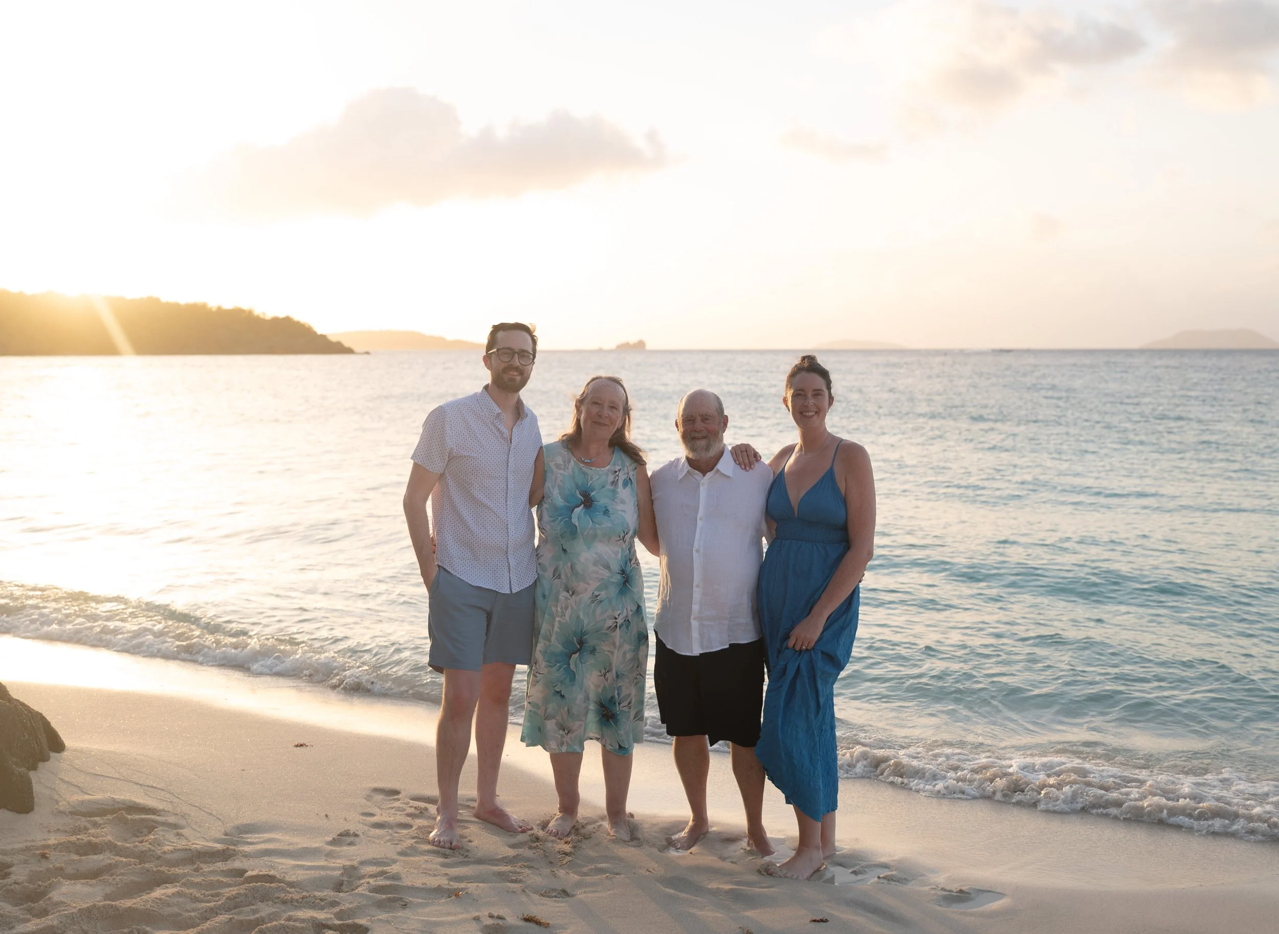 Four people standing together on a sandy beach during sunset, with the ocean and distant land in the background. The group includes two men and two women, all smiling at the camera.