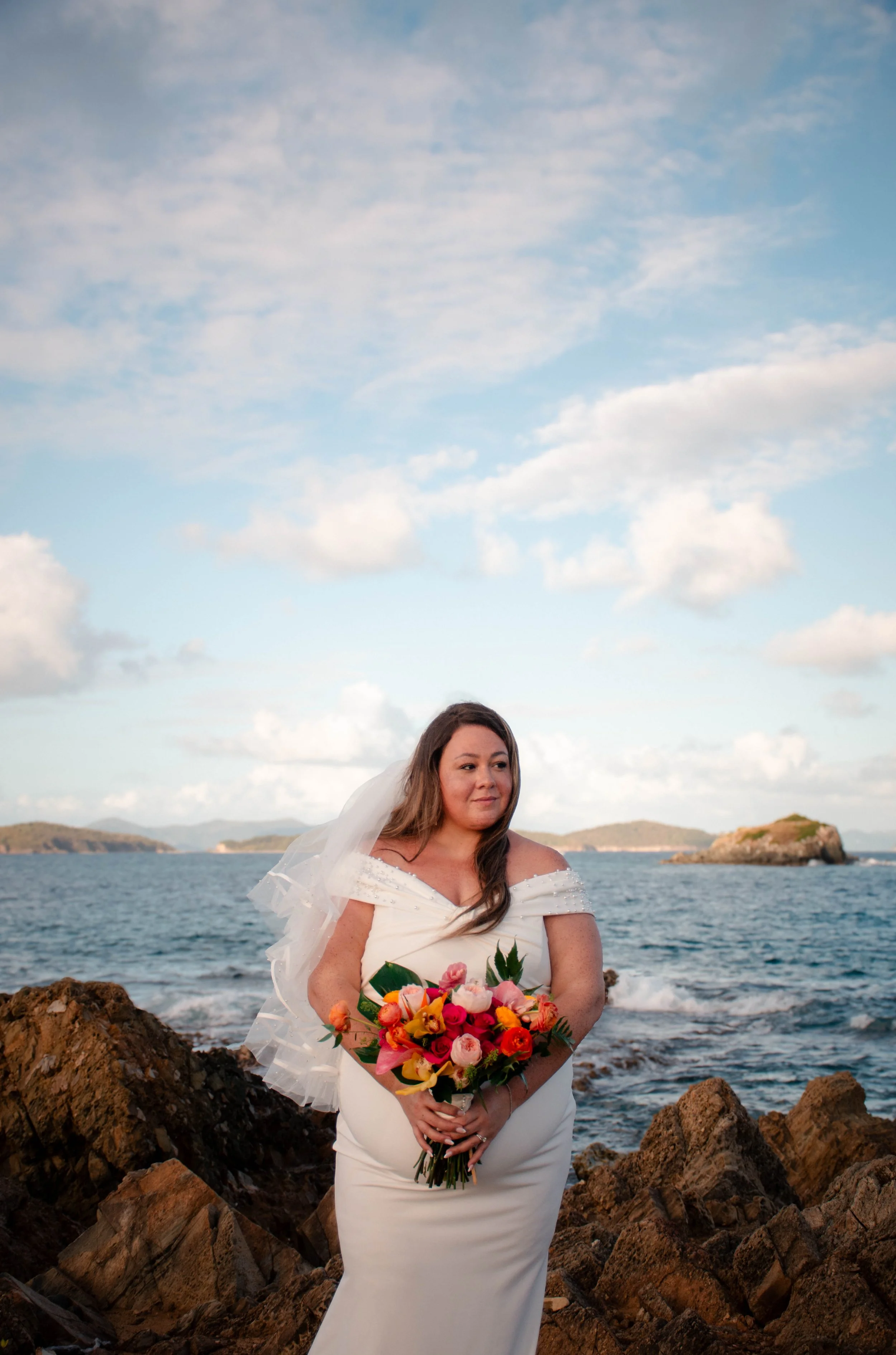 A bride in a white wedding dress holding a bouquet of colorful flowers on a rocky shoreline with the ocean and islands in the background, under a partly cloudy sky.