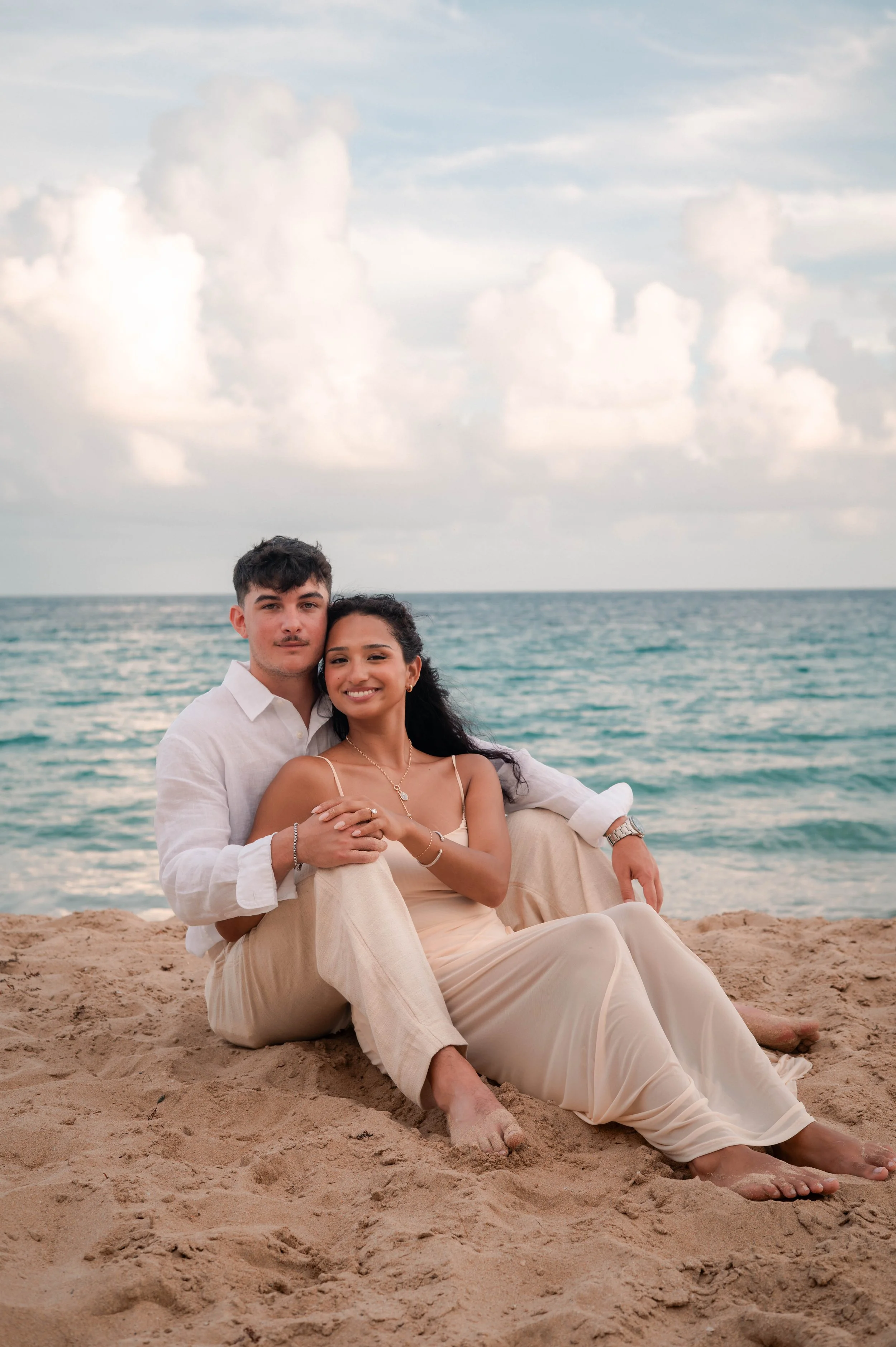 A young couple sitting on the sandy beach near the ocean, with the man embracing the woman. Both are smiling, dressed in light-colored casual clothing, with the ocean and cloudy sky in the background.