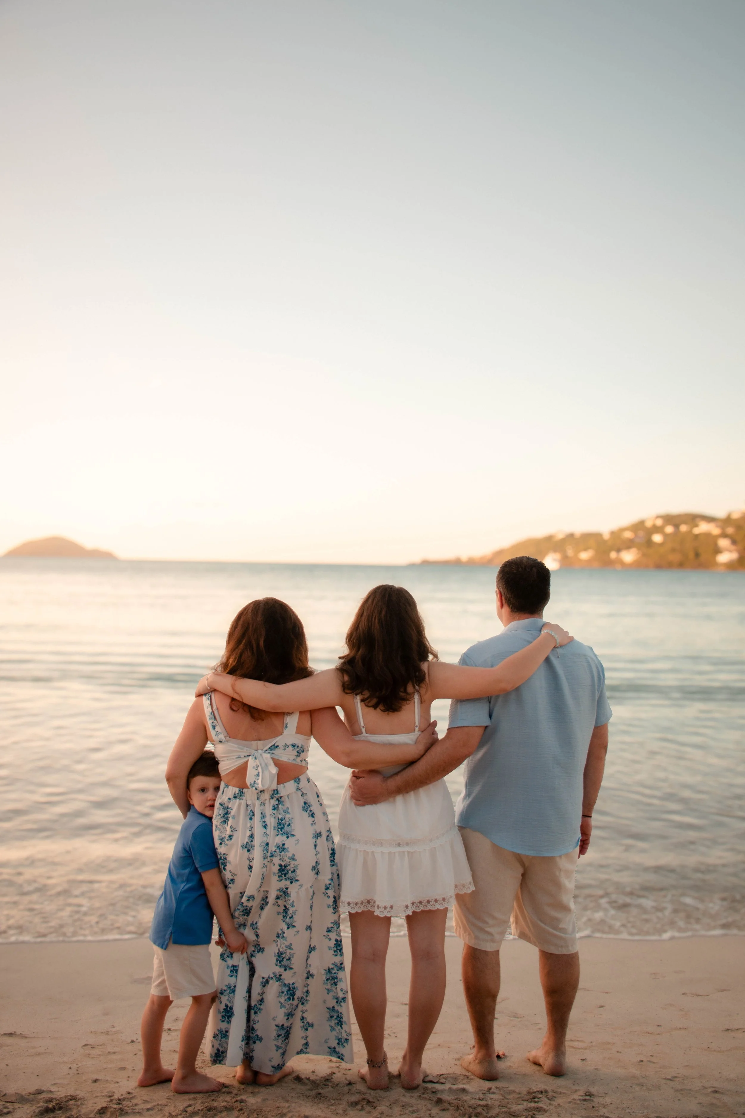 A group of five people, three women, one man, and one young boy, are standing on a beach facing the water and sunset. The women are hugging and the young boy is standing at their feet, with a backdrop of hills, calm water, and a clear sky.
