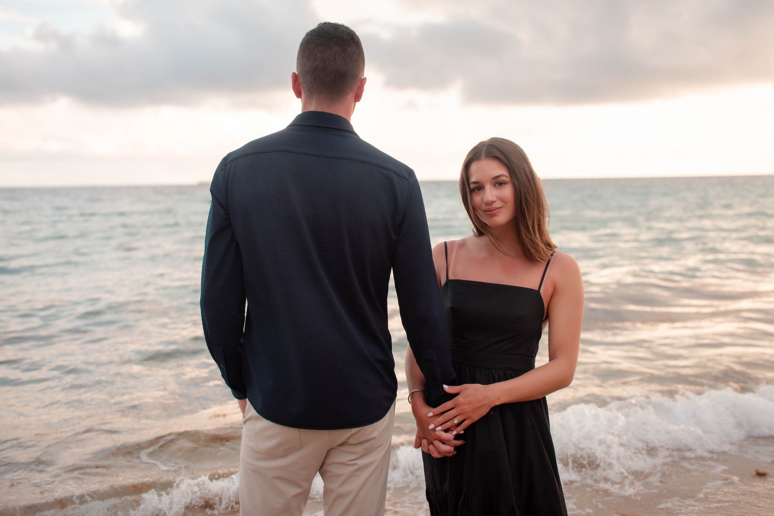 A man and woman standing on a beach, holding hands, with the ocean and cloudy sky in the background.